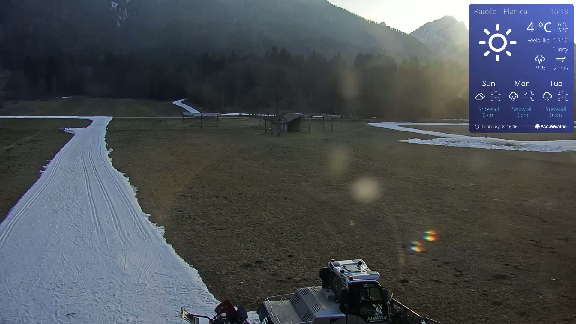 A snow groomer sits near a prepared cross-country ski trail on a sunny day in a field, with mountains in the background.