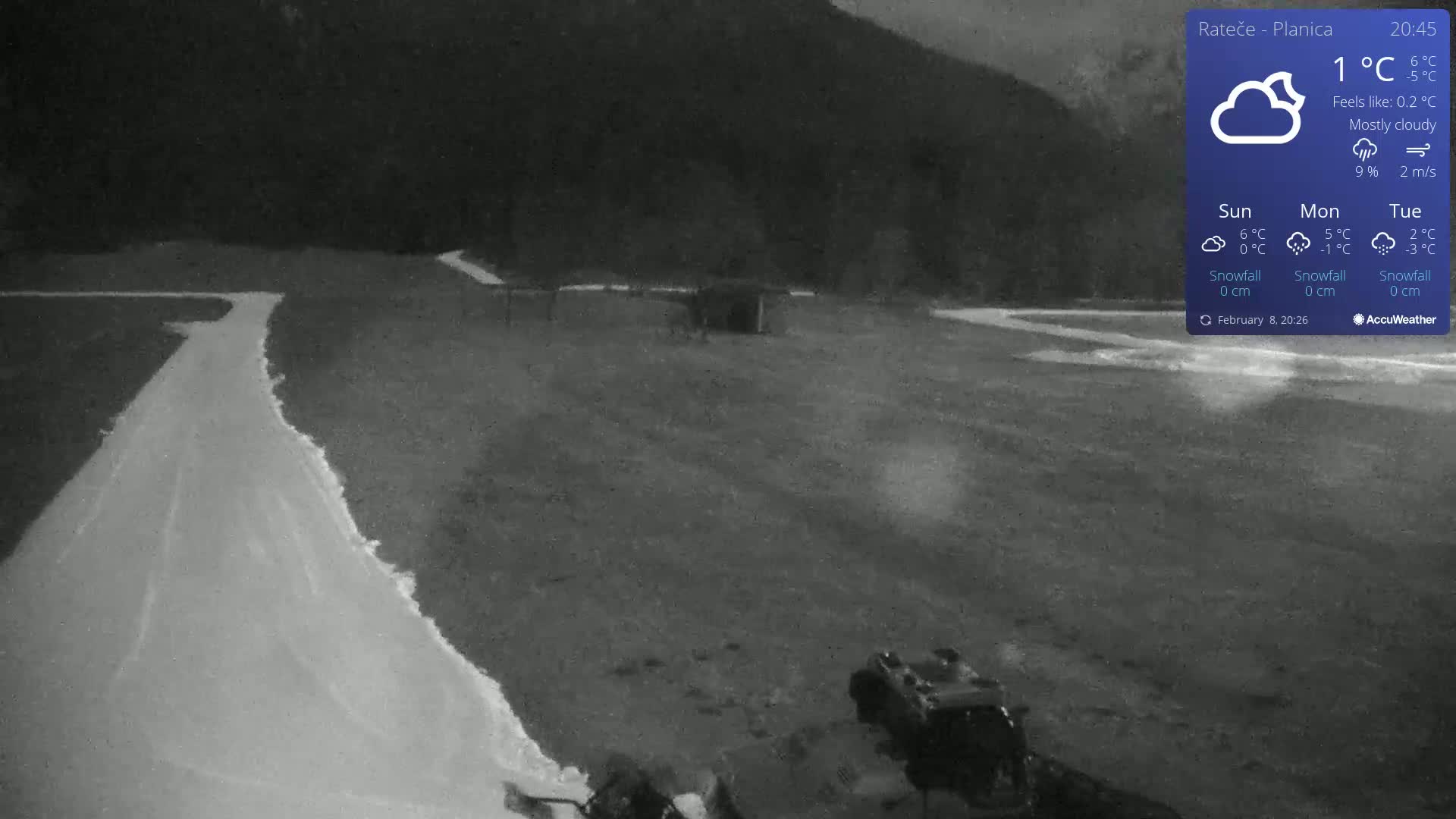 A mostly cloudy night scene shows a snow-covered path winding through a dark field, with a small structure and machinery visible in the distance.