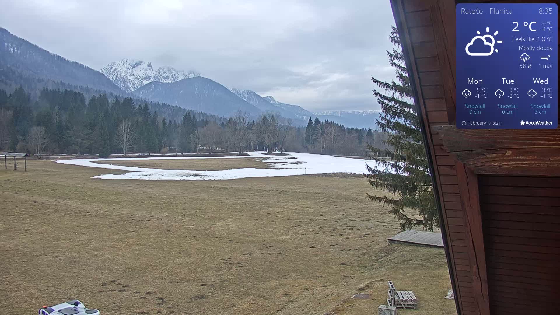 A mostly cloudy, 2°C day reveals a snow-dusted field and distant mountains, partially covered in snow, viewed from a wooden structure.