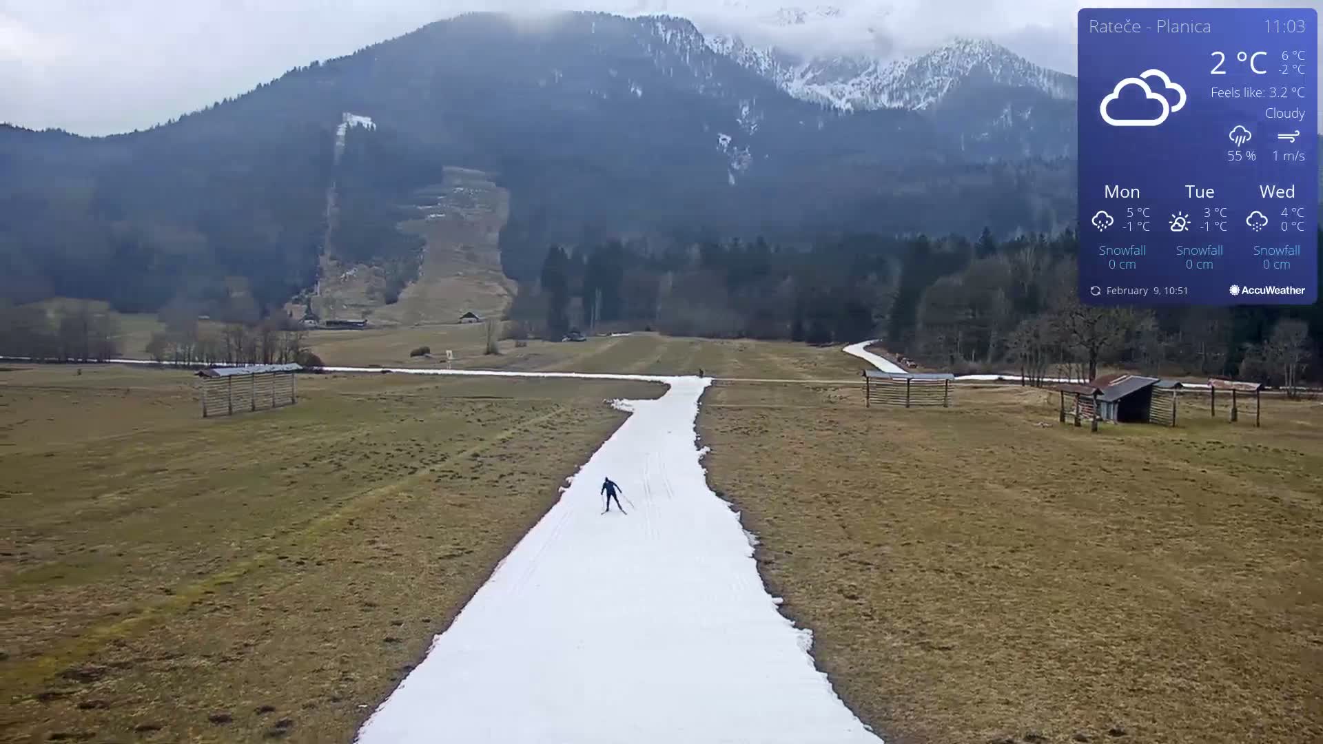 A cross-country skier is on a snow-covered path in a grassy field, with mountains in the background under cloudy skies.