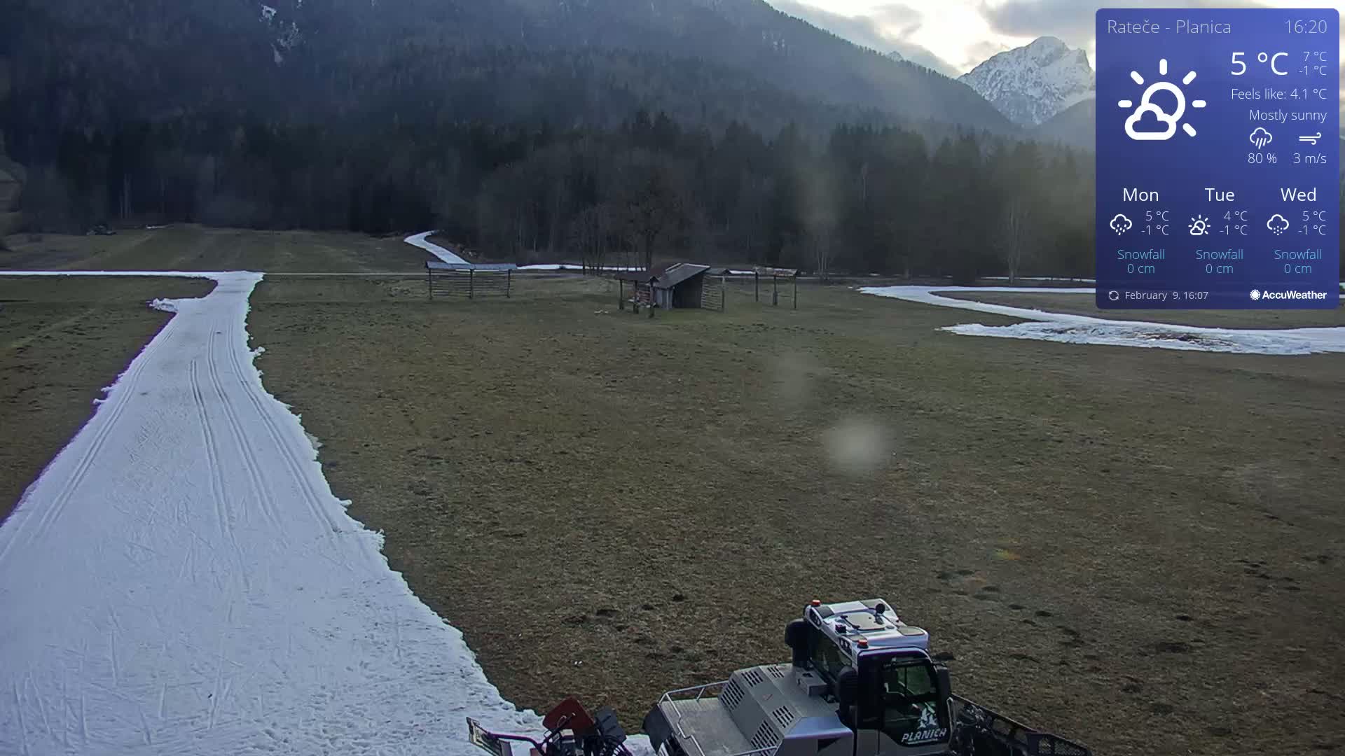 A snow groomer is parked on a mostly brown field with a narrow snow-covered path winding through it, under a mostly sunny sky in front of a mountainous backdrop.