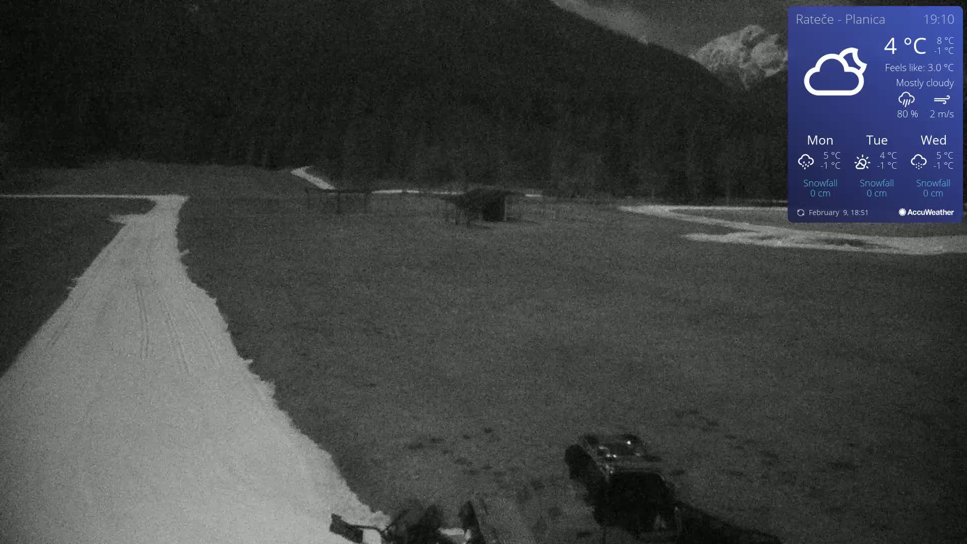 A mostly cloudy night scene shows a snow-covered path winding through a dark, flat area, with a vehicle and some equipment visible in the lower right corner, and a mountainous backdrop.
