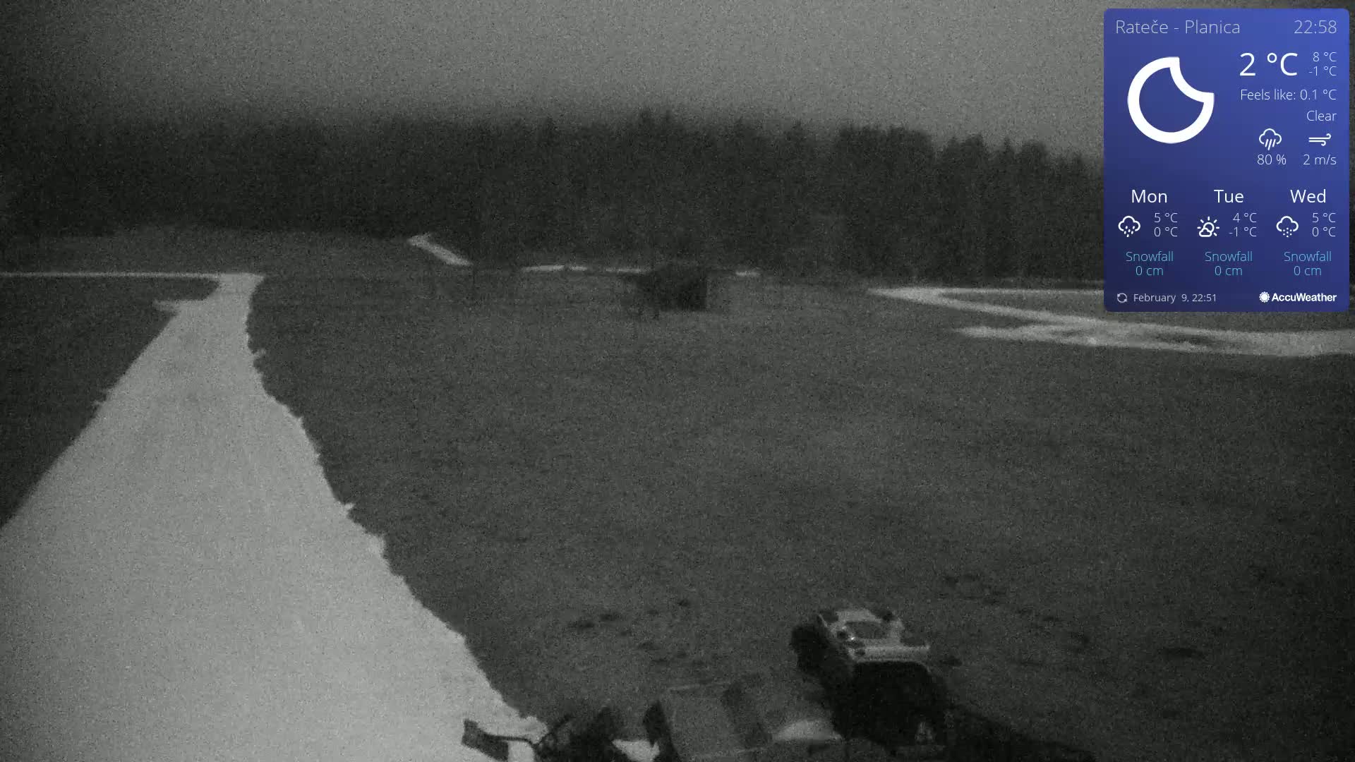 A nighttime, clear view of a snow-covered area with vehicles and a dark treeline in the background.