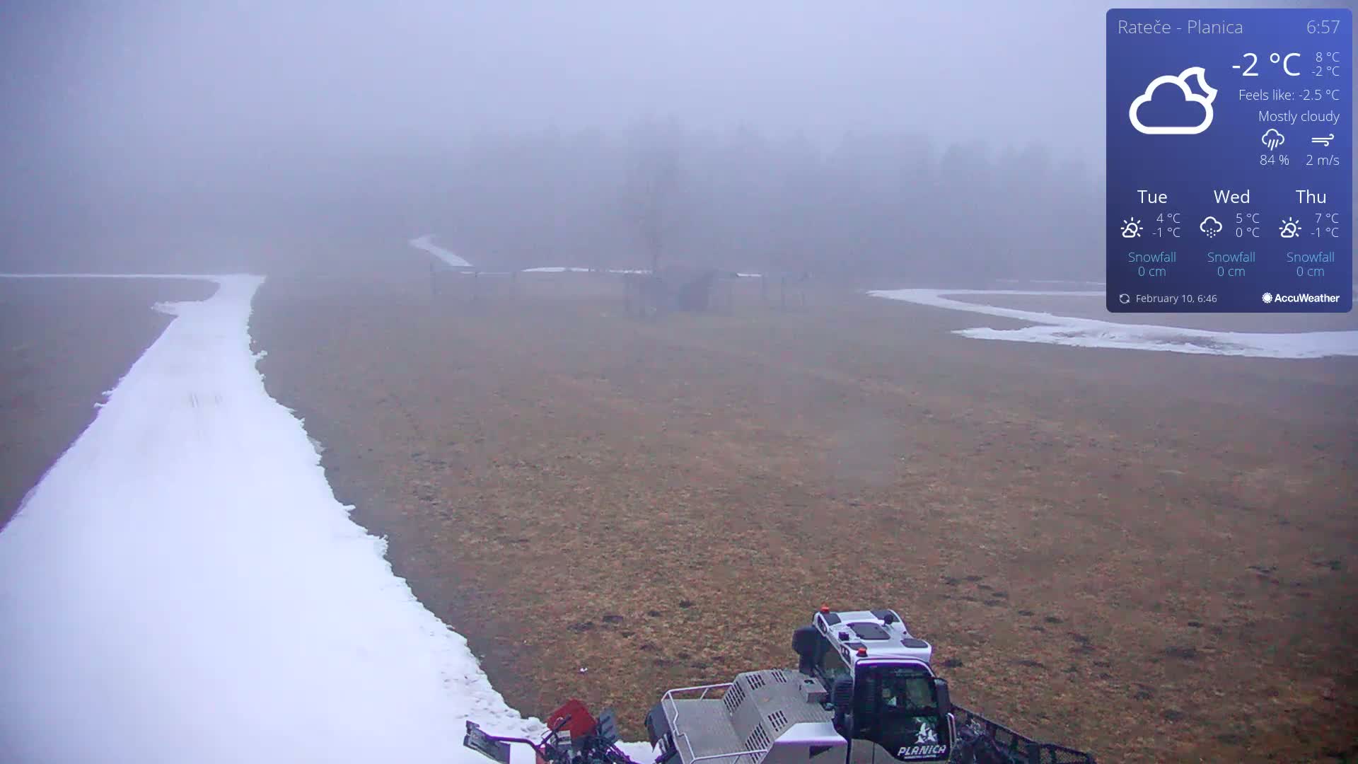 A snow groomer is partially visible at the bottom of the image, working on a snow-covered path that cuts through a mostly brown, foggy field.
