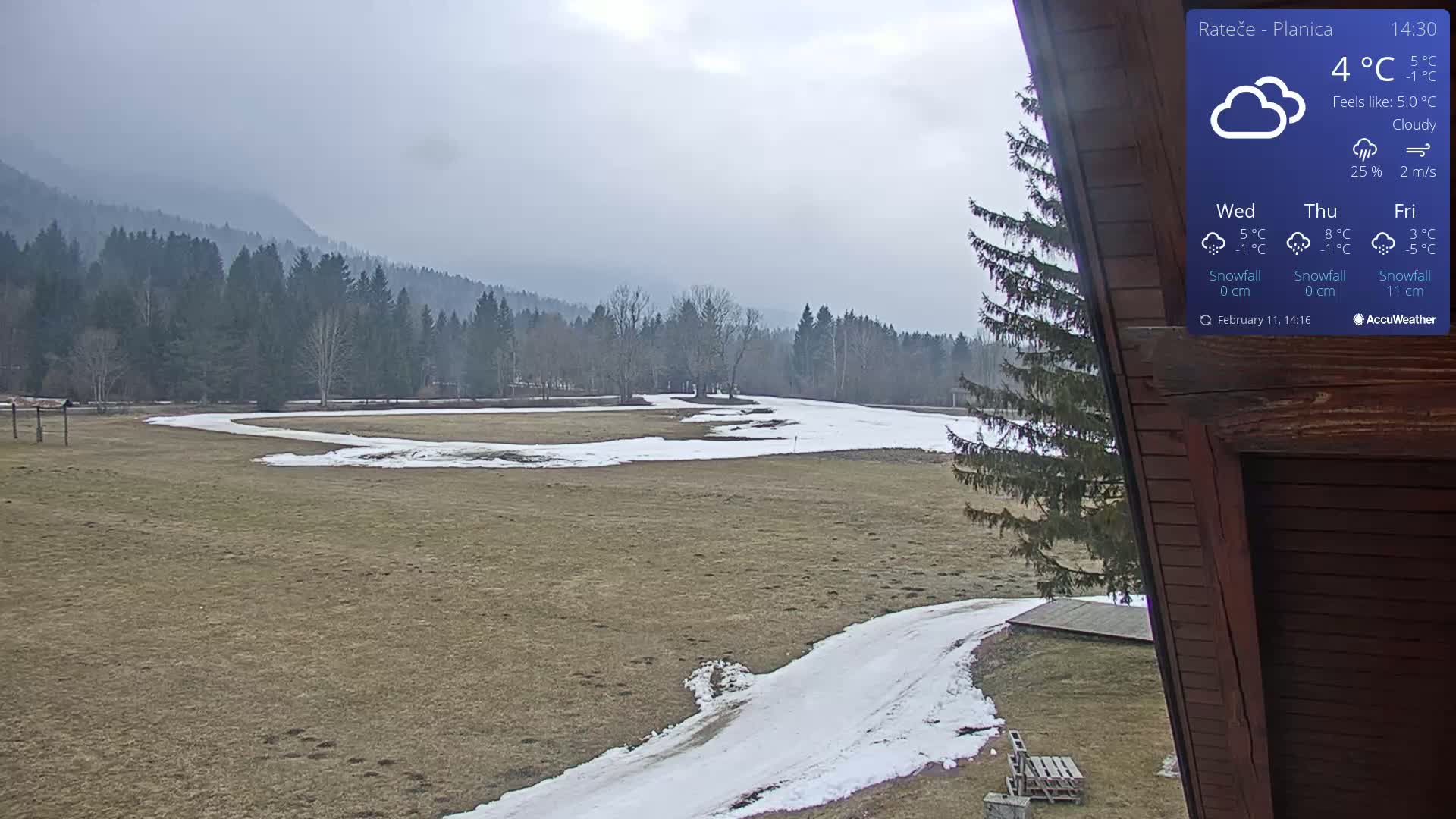 A cloudy, outdoor view shows a partially snow-covered field with a winding path leading toward a line of dark evergreen trees and mountains in the distance.
