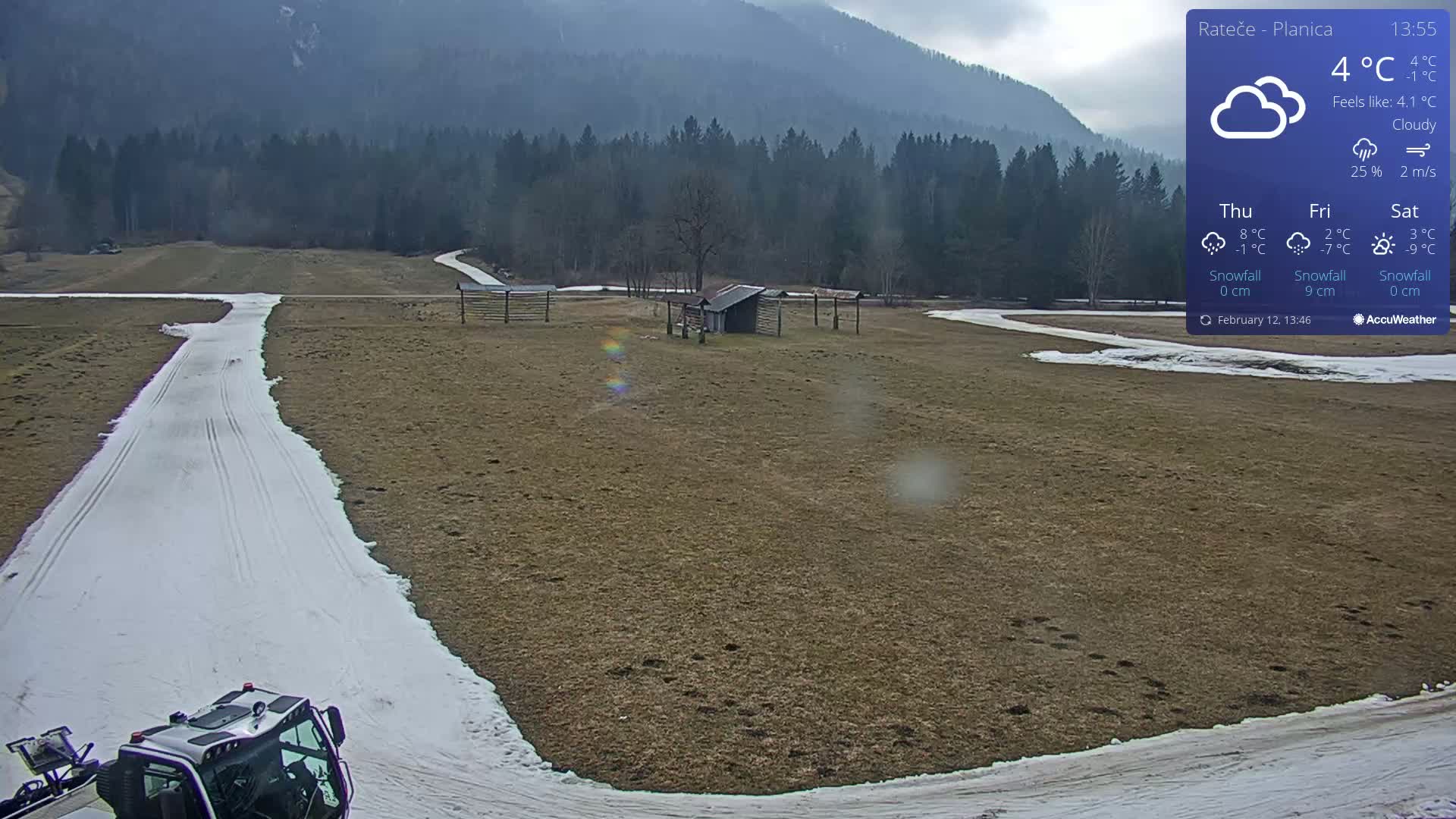 A snow-grooming machine is parked on a snow-covered track in a field, with a cloudy sky and mountains in the background.