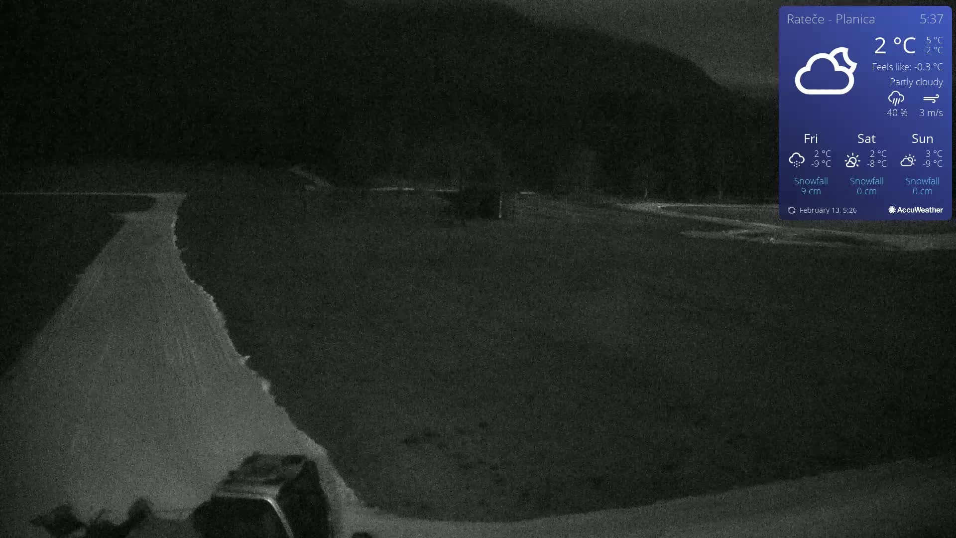 A dark, partly cloudy night scene shows a snow-covered road curving into a flat, dark area under a hill.