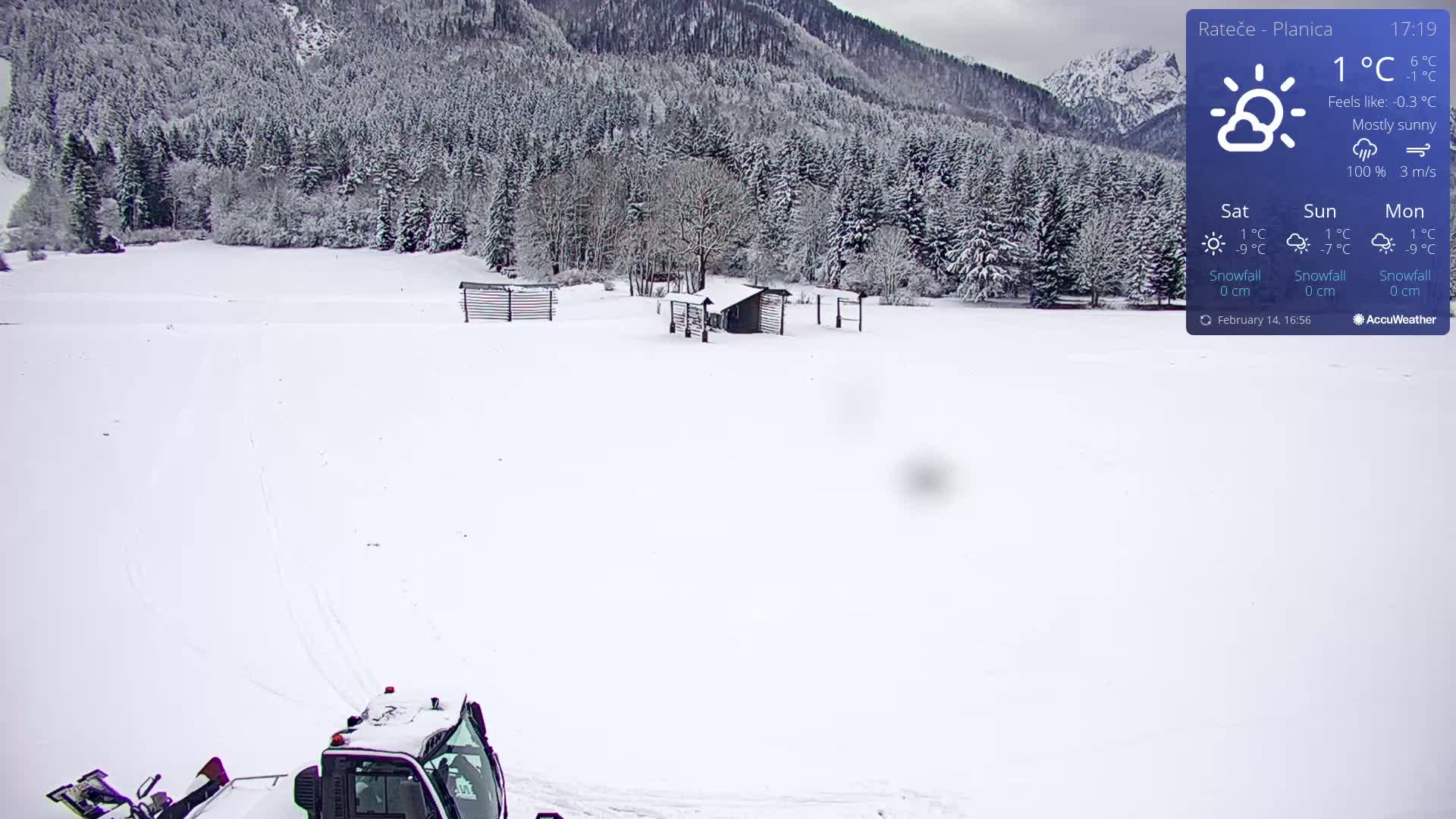 A snow-covered field with a snowcat in the foreground, nestled amongst snow-laden trees and a mountain backdrop under mostly sunny skies.