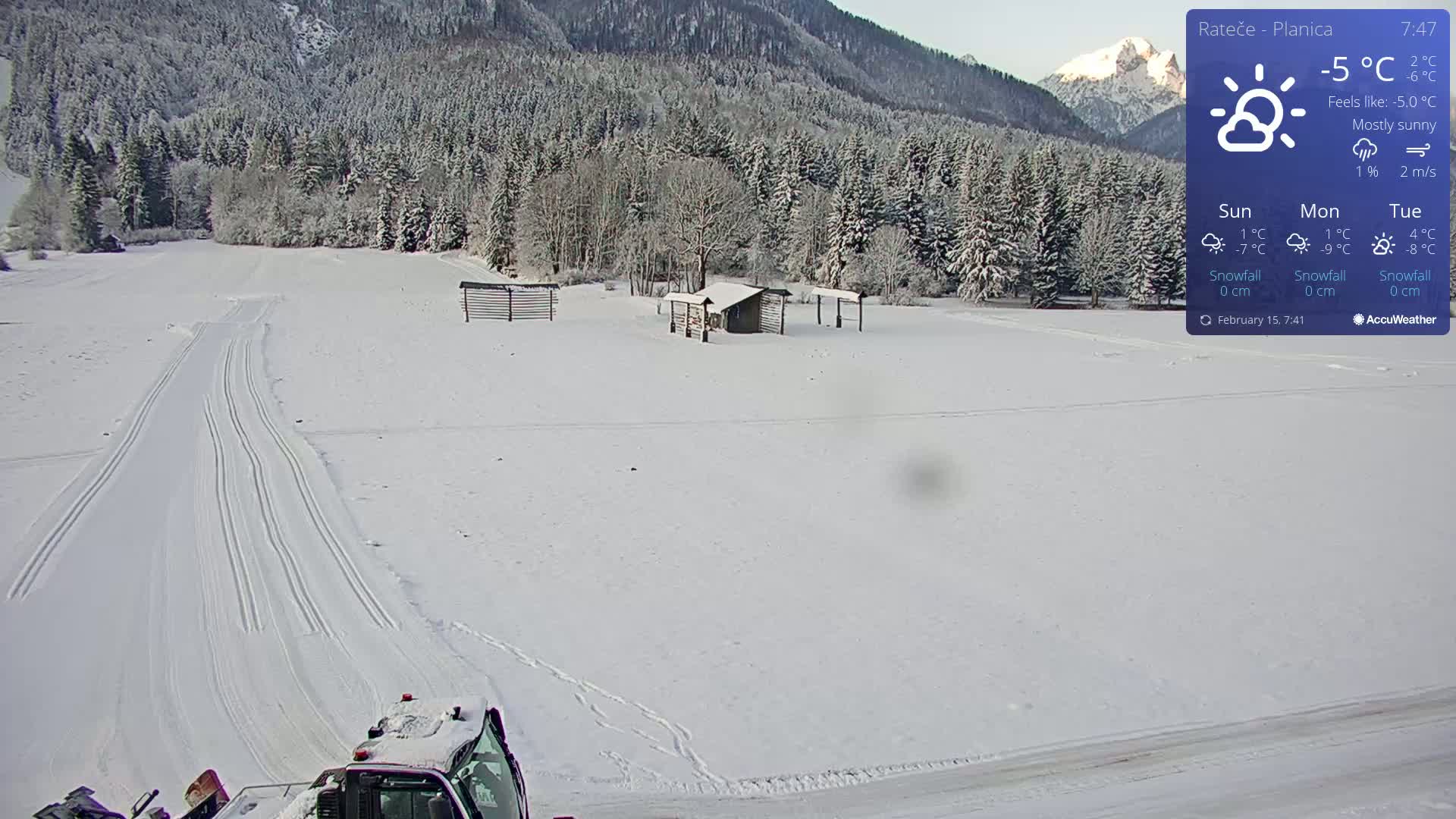 A snow-covered landscape with groomed cross-country ski trails, a small building, and a snow groomer in the foreground under mostly sunny skies.