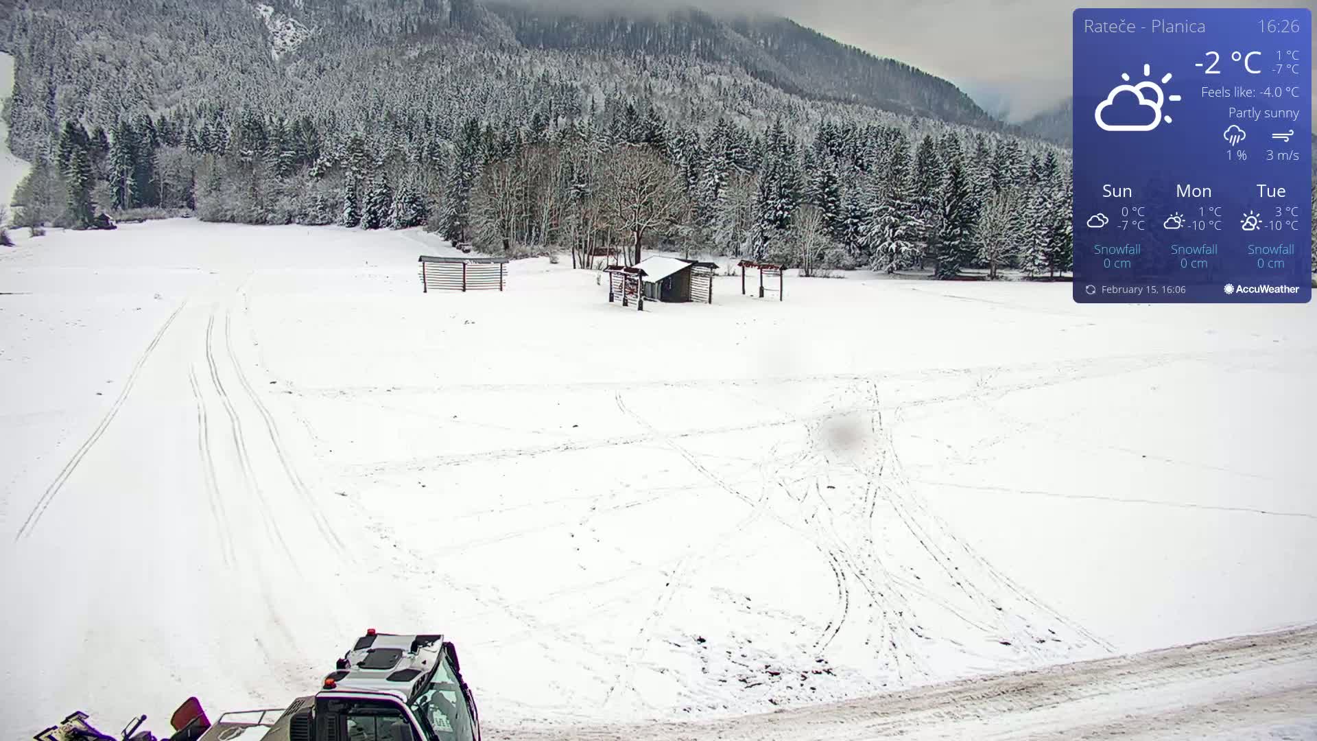 A snow-covered field with tire tracks and a small building is visible, with a partly sunny sky and a snow-grooming machine in the foreground, backed by a snow-covered forest and mountains.