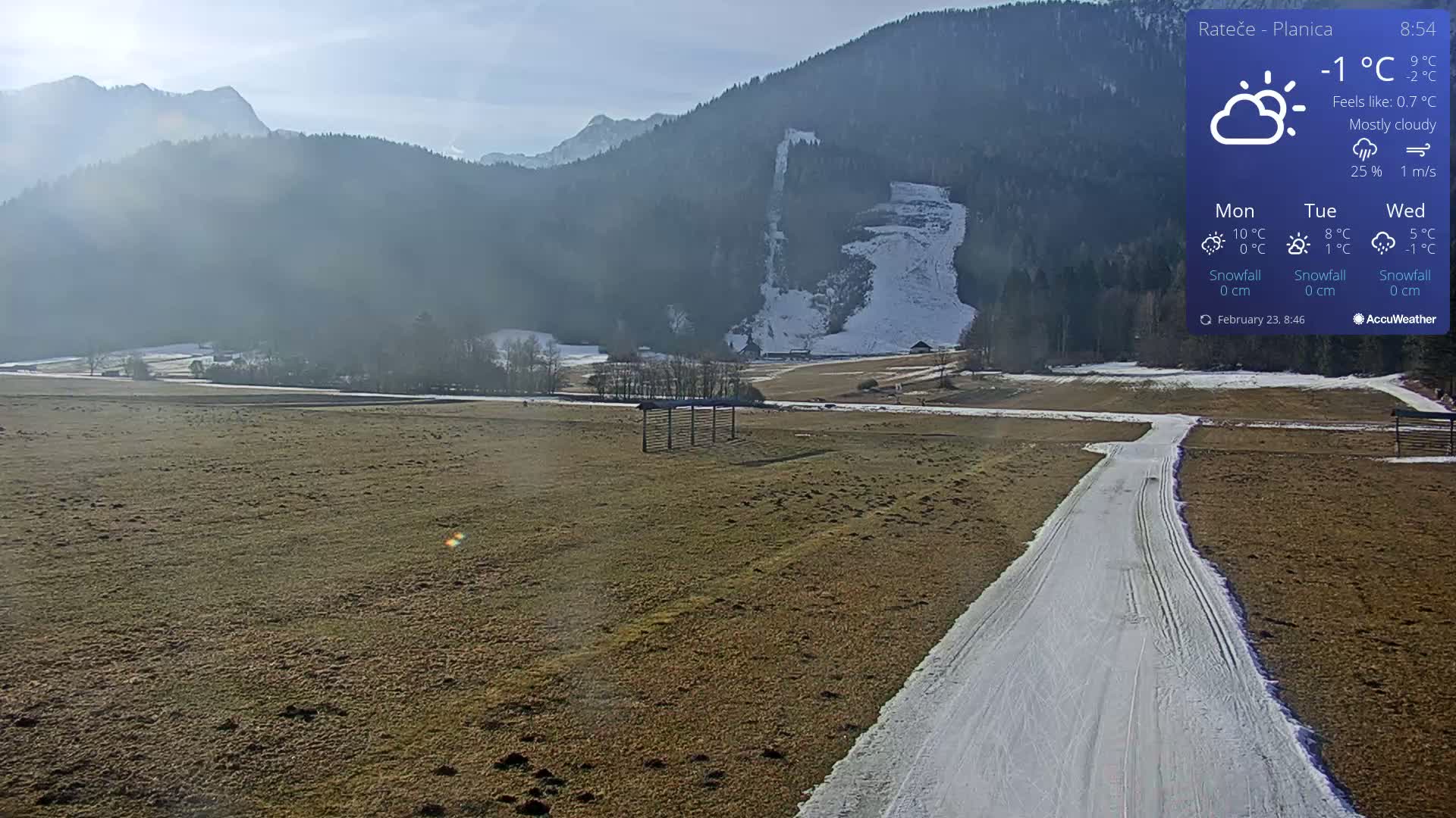 A mostly cloudy, cold day shows a snowy mountainside in the background, with a flat, snow-covered path winding through a brown field in the foreground.