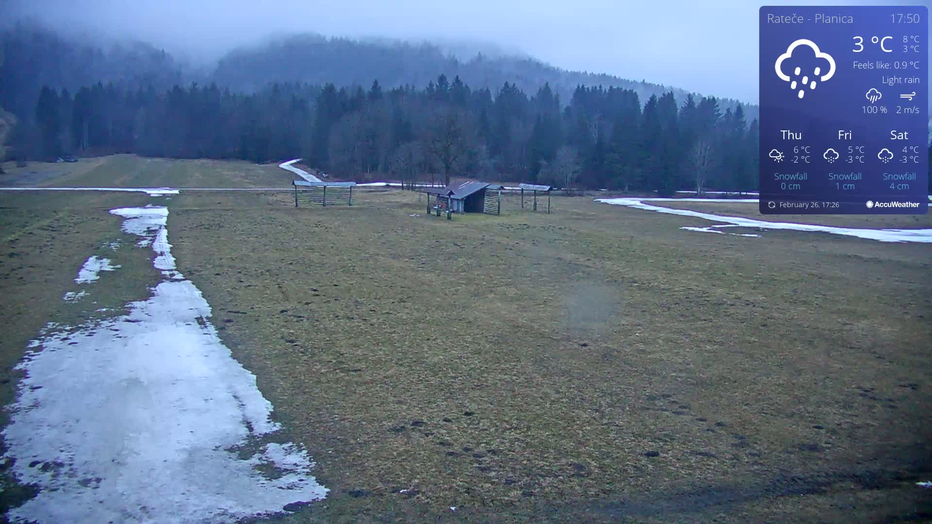 A flat, grassy field with patches of snow and ice under a light rain, shows a small shed and wooden structures, with a dark, foggy forest in the background.