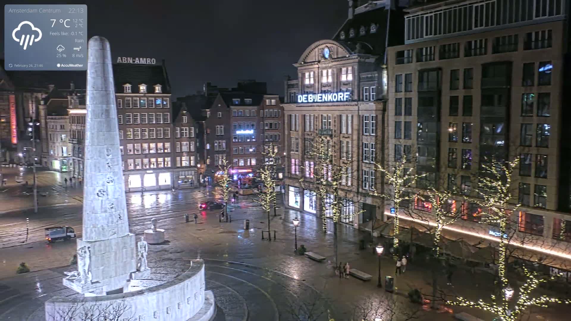 A nighttime, rainy view of a city square, centered on a tall monument, with illuminated buildings and trees decorated with lights.