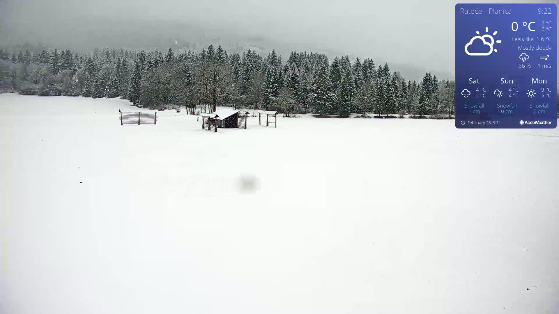 A snow-covered field with a small cabin and fence, backed by a snow-dusted evergreen forest under a mostly cloudy sky.
