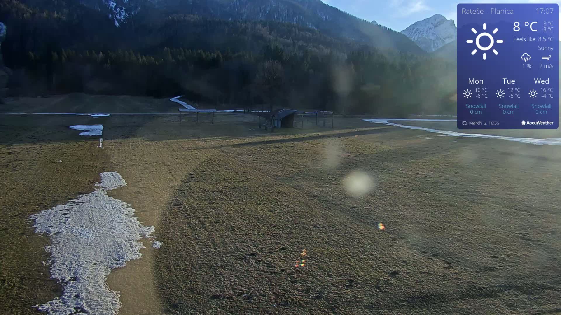 A sunny, mostly clear day reveals a field with patches of snow, a small structure, and a mountain range in the background.