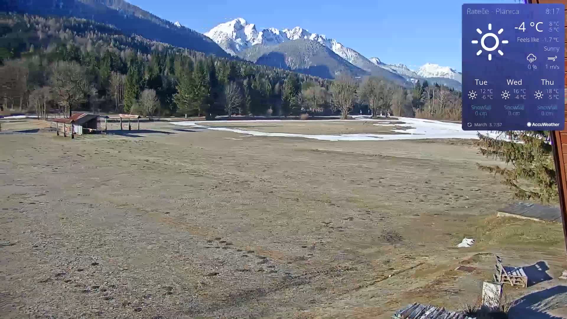A sunny day reveals a flat, barren landscape with a small wooden structure, a background of coniferous forest, and snow-capped mountains in the distance.