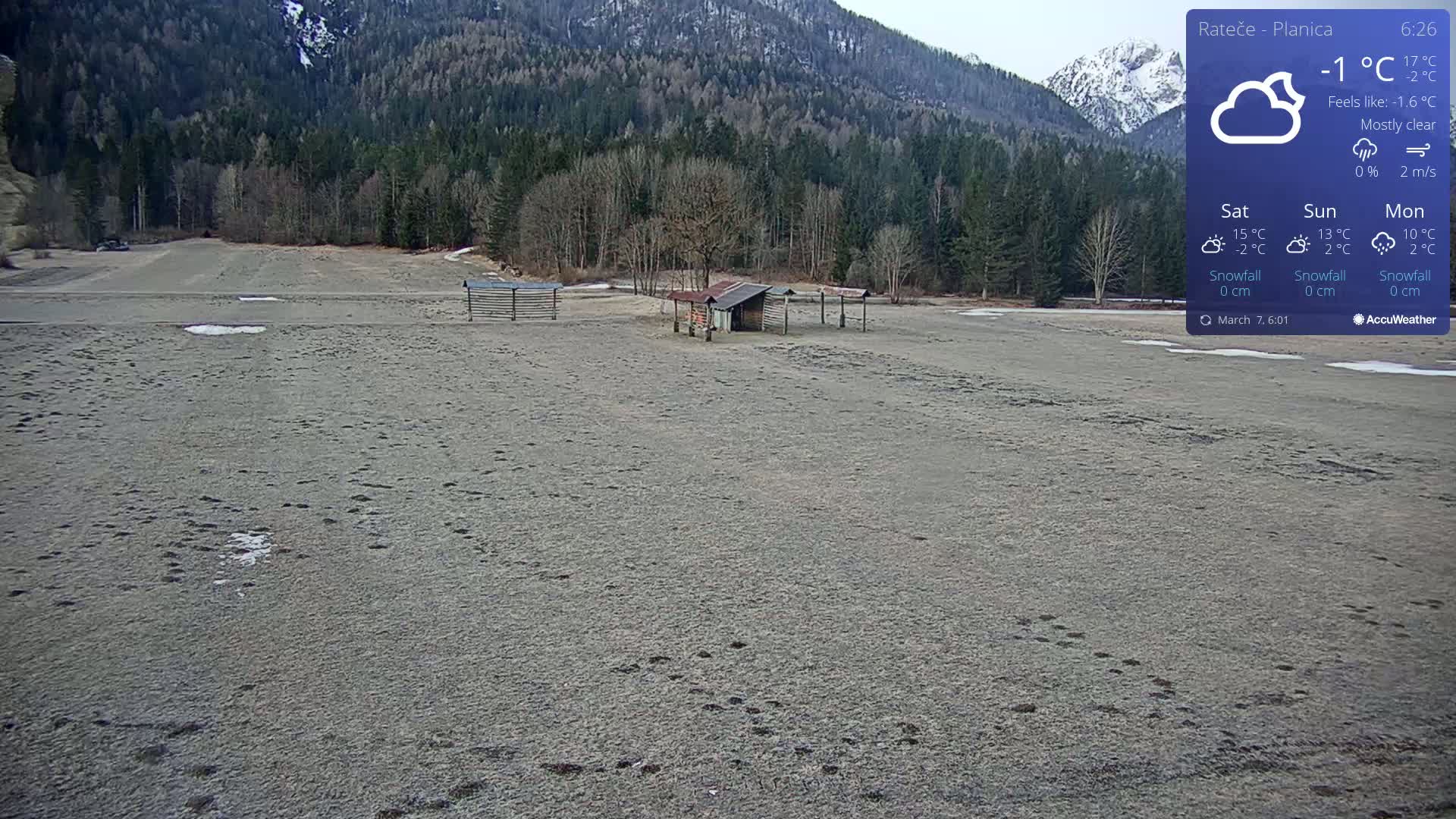 A mostly clear, cold day with a temperature of -1°C shows a flat, snow-dusted field with animal tracks leading to a small shed, backed by a forest and mountains.