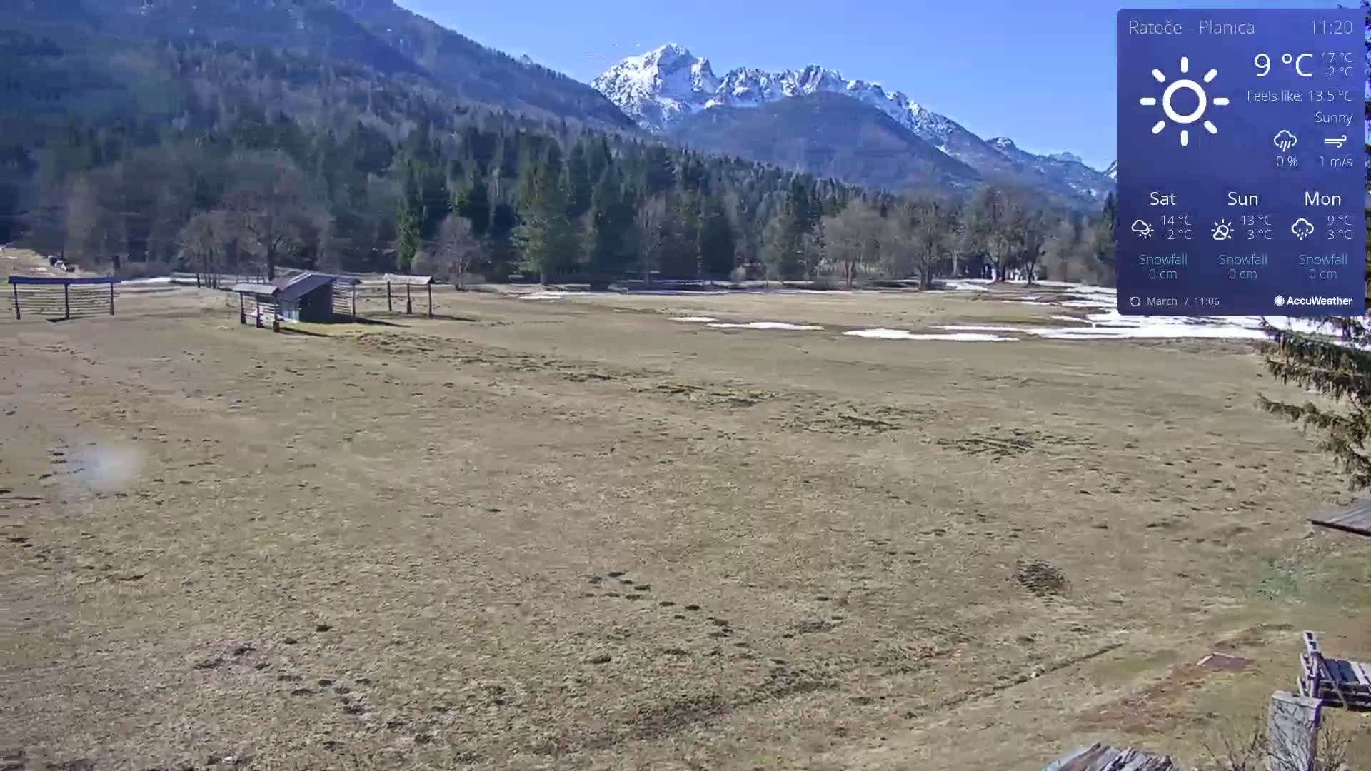 A sunny, open field with a small shed and wooden fencing is situated in front of a snow-capped mountain range and a forest, under a clear blue sky.