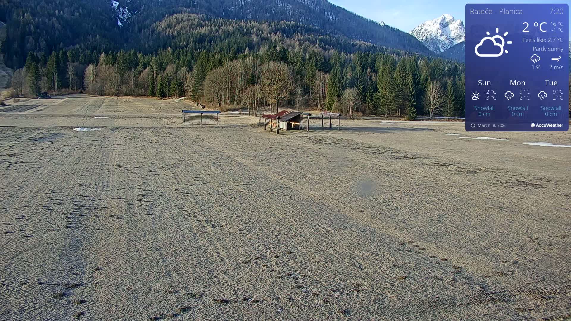 A mostly sunny, sparsely populated, sandy field with a small structure and fencing is situated before a background of a coniferous forest and snow-capped mountains.