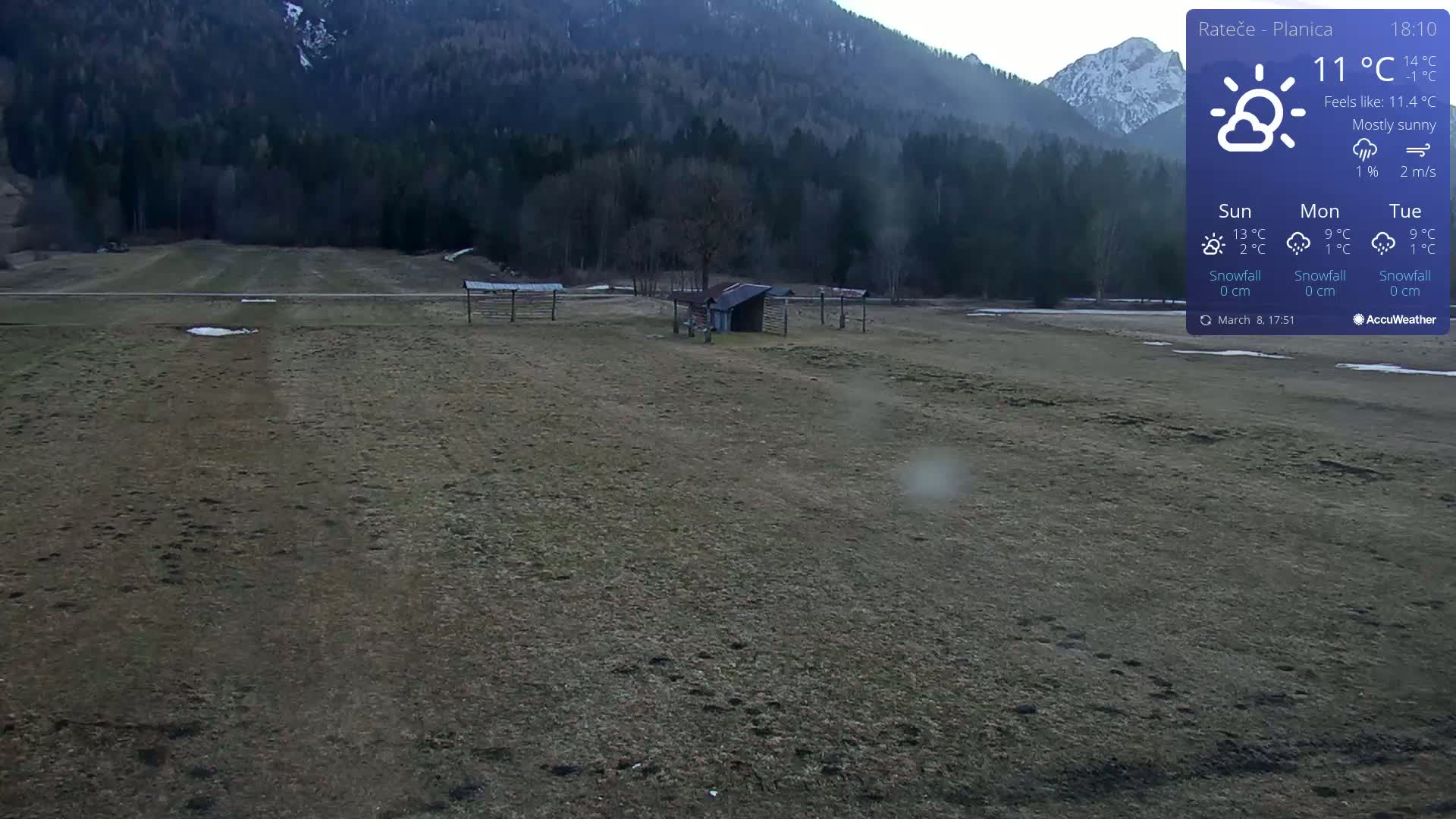 A mostly sunny, flat, brown field with a small shed and wooden structures, backed by a mountain range under a mostly clear sky.