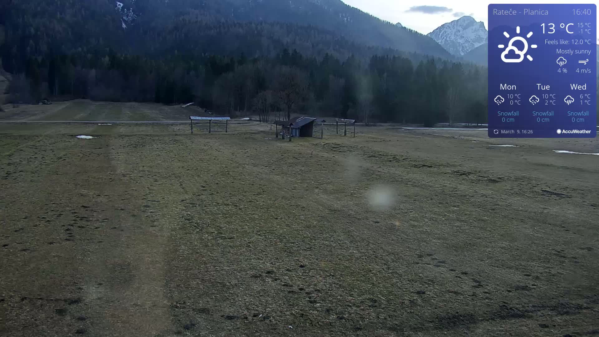 A mostly sunny, flat, grassy field with a small shed and wooden structures sits before a mountain range under a mostly clear sky.