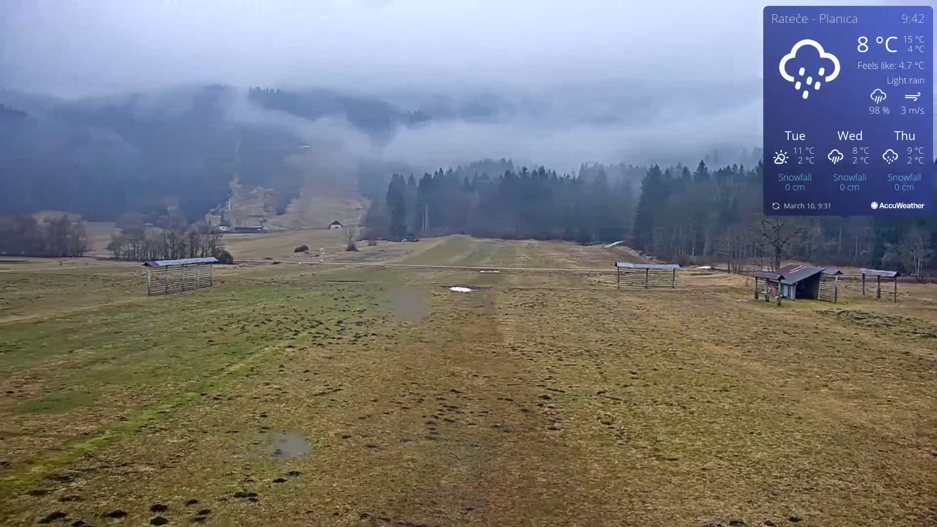 A flat, grassy field with a few small structures is visible in the foreground, with misty mountains and a forest in the background under light rain.
