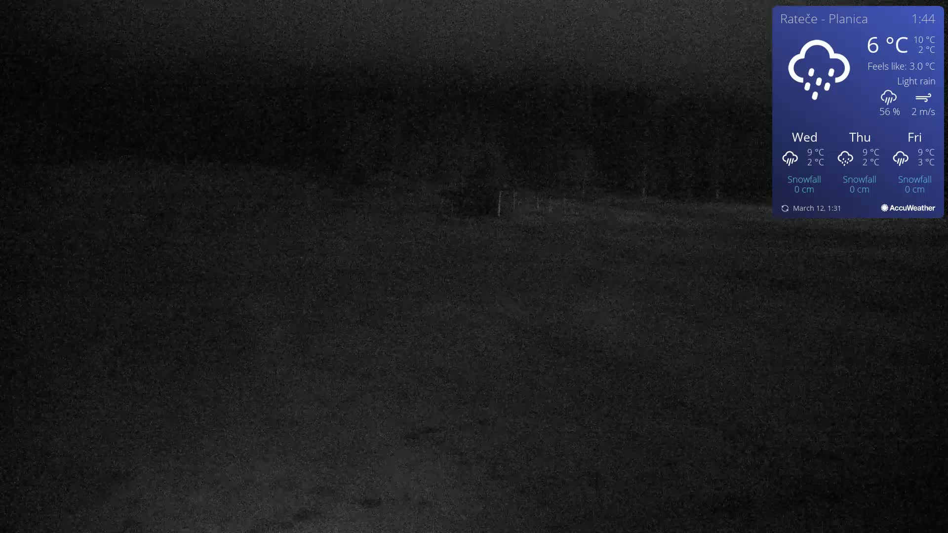 A dark, nighttime outdoor scene shows light rain falling on a flat, dark-colored area.
