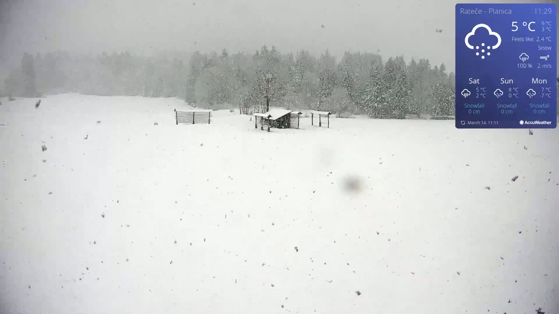 A snow-covered field with a small wooden structure and fences is seen during a snowfall.