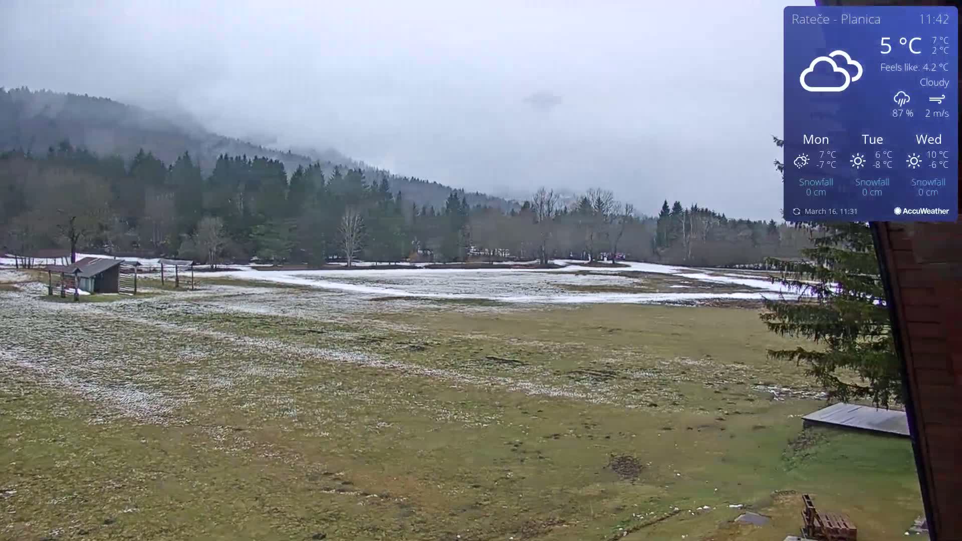 A snow-dusted field stretches out before a line of dark green trees and misty mountains under a cloudy sky.