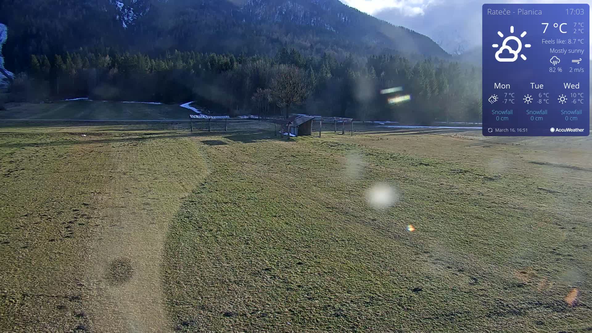A mostly sunny, outdoor view shows a flat, grassy field with a small shed and distant mountains under a mostly sunny sky.