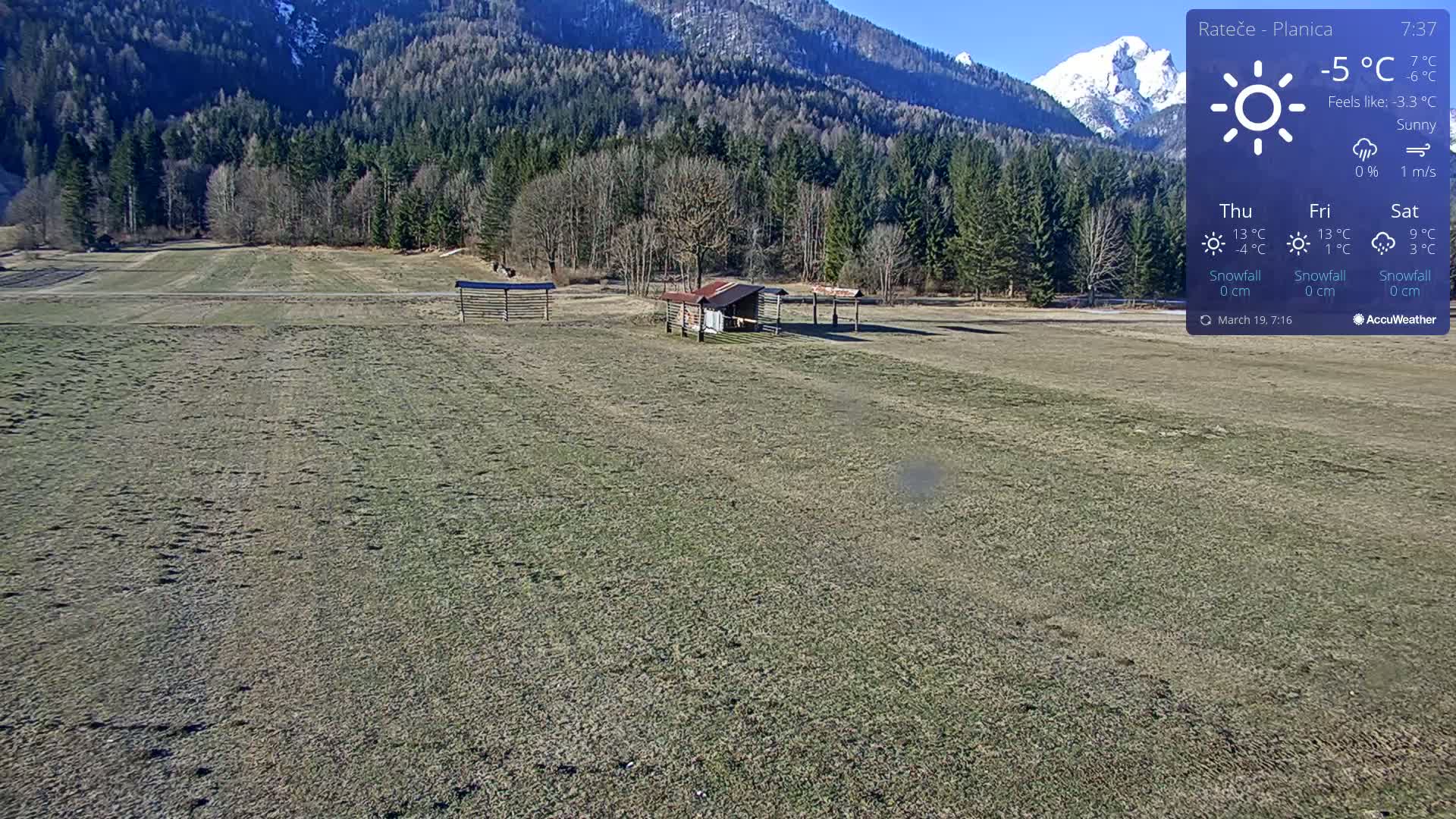 A flat, grassy field with a small shed and wooden structures sits before a background of dark green forested hills and snowy mountains under a sunny sky.