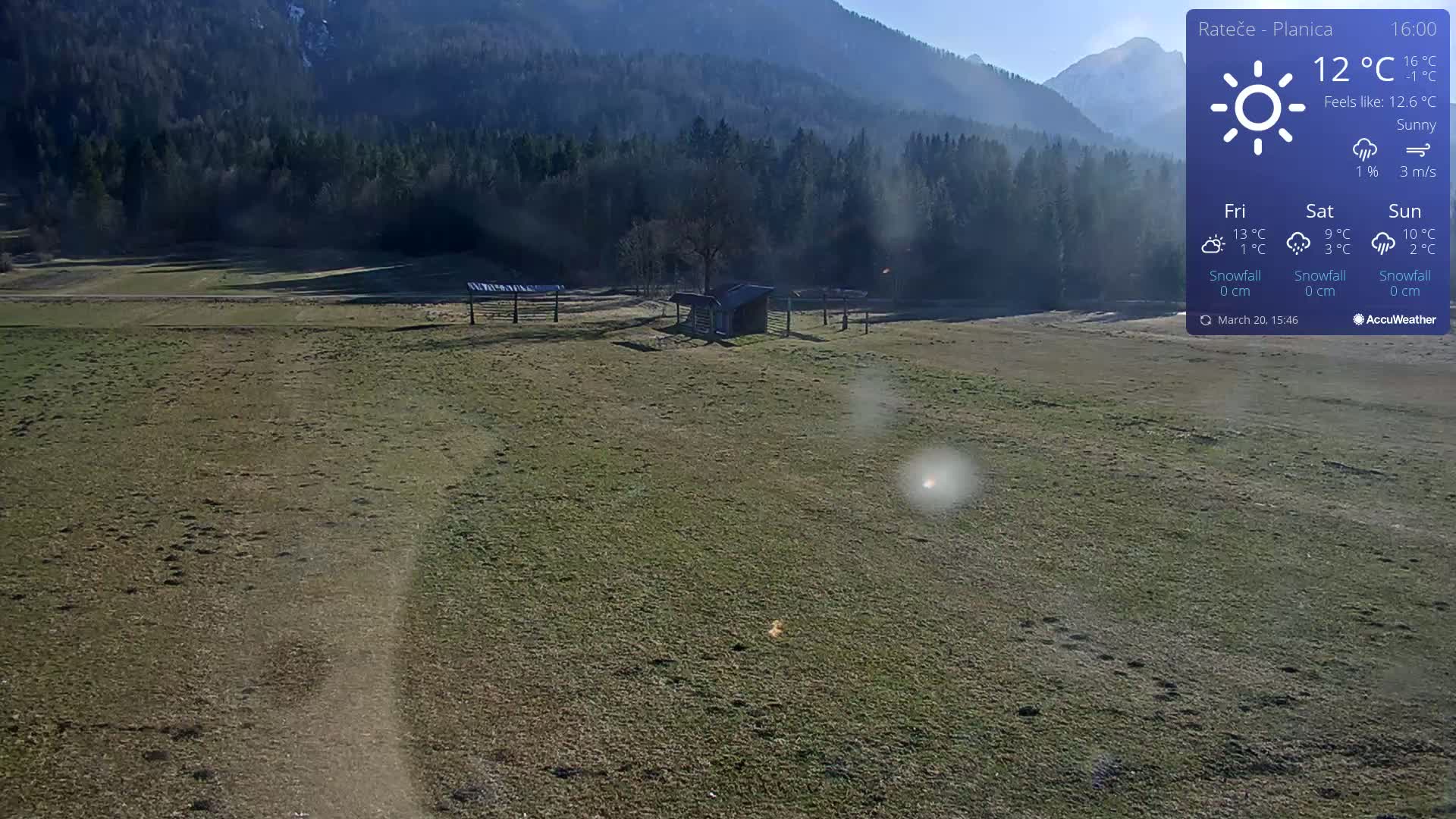 A sunny, grassy field with a small shed and some fences stretches out before a background of dark green mountains.