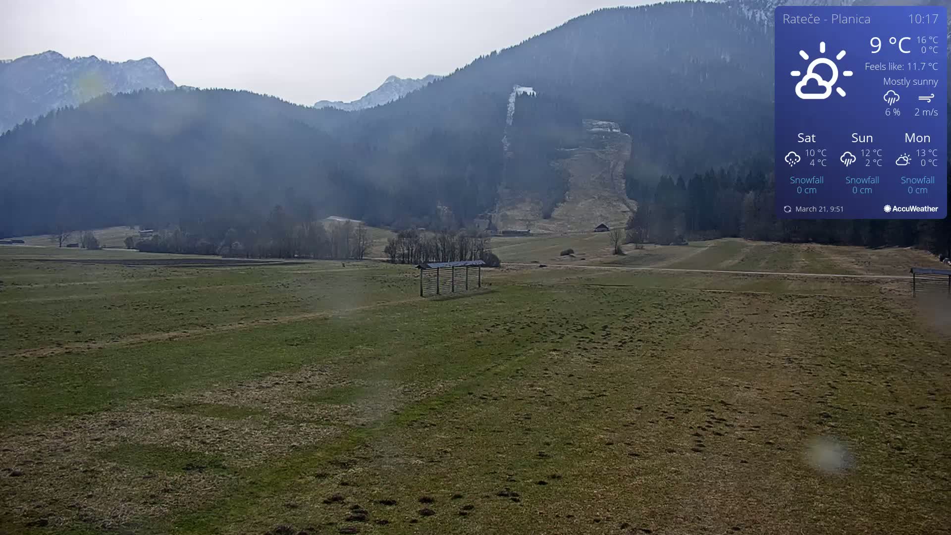 A mostly sunny, flat, grassy field stretches out before a backdrop of dark, tree-covered mountains under a pale sky.