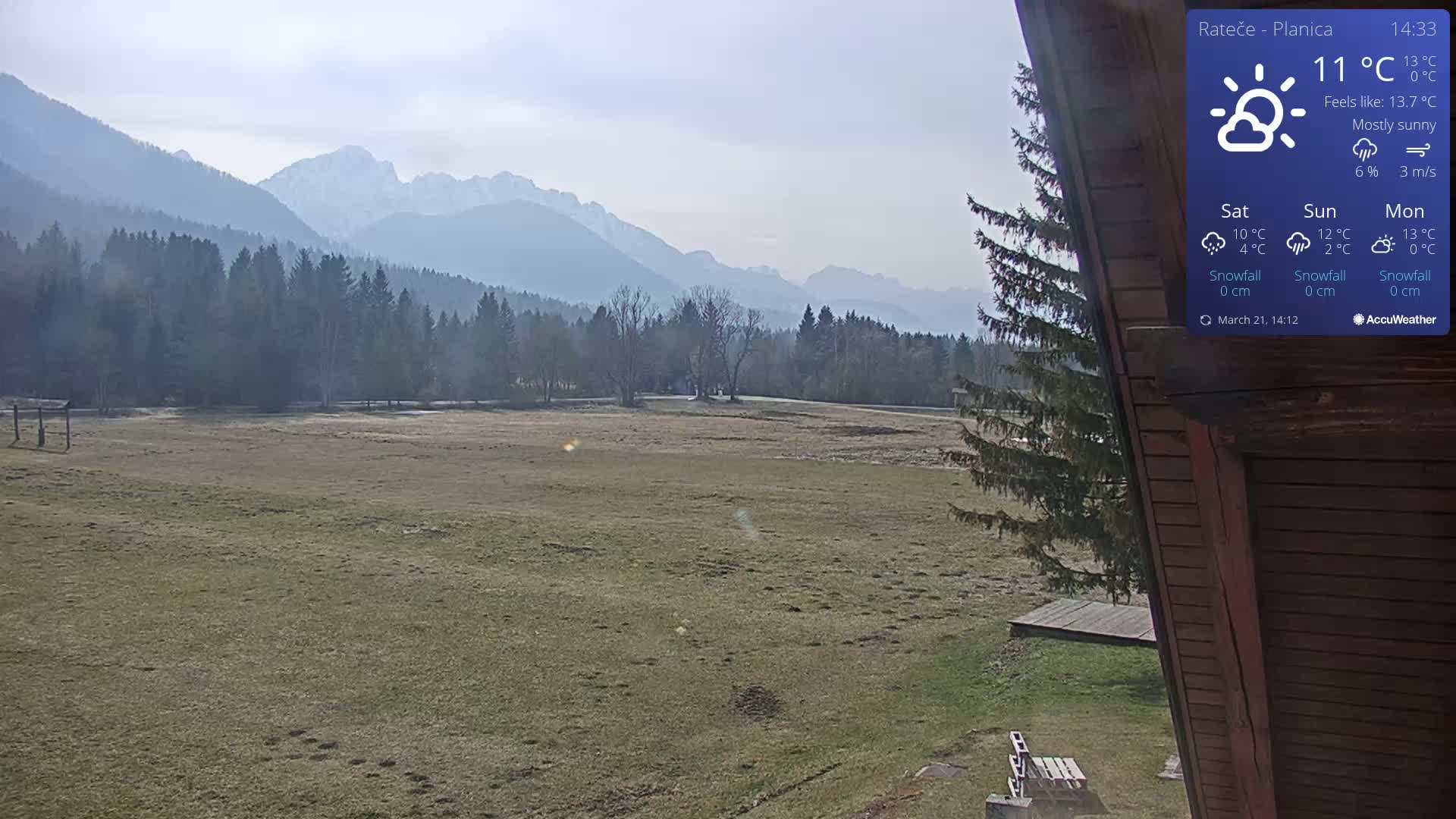 A mostly sunny view of a grassy field and dark green forest with snow-capped mountains in the background.