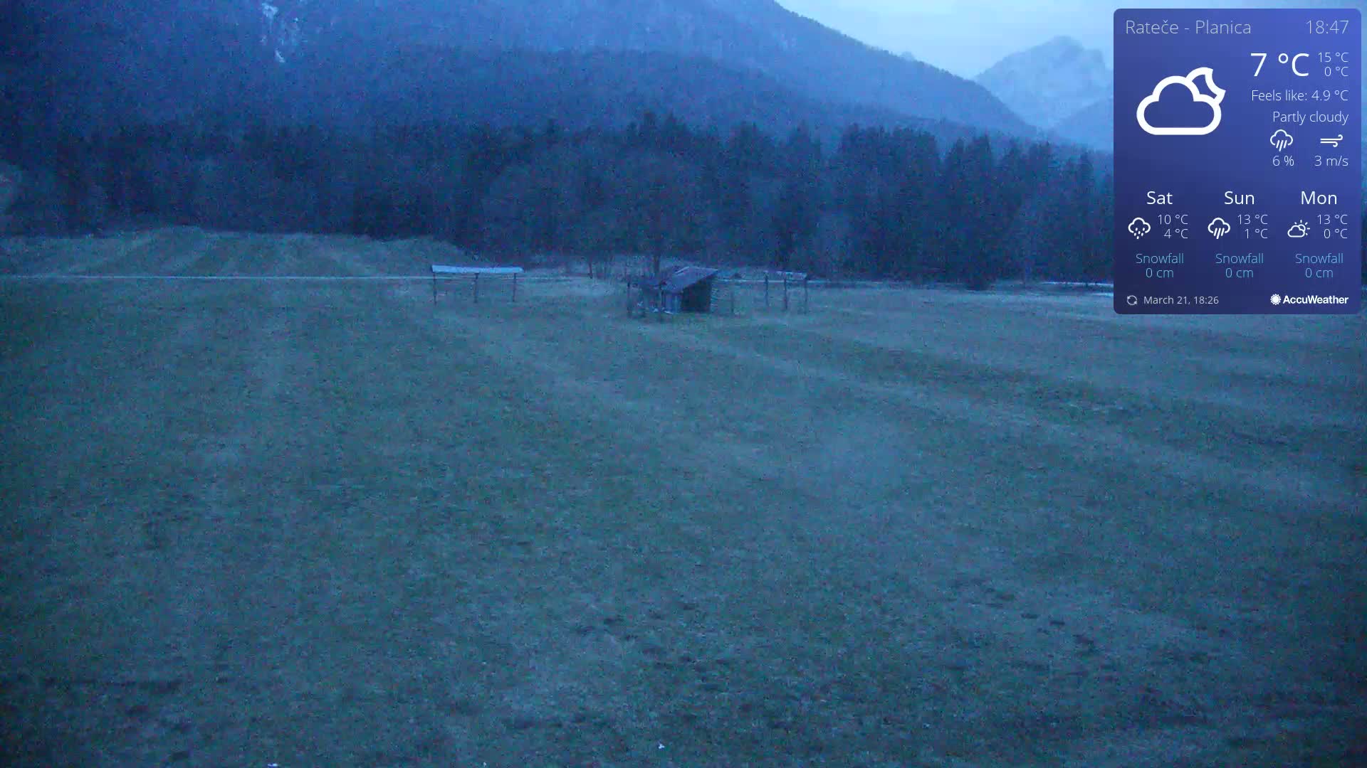 A flat, grassy field with a small shed sits beneath a partly cloudy, twilight sky in front of a mountain range.