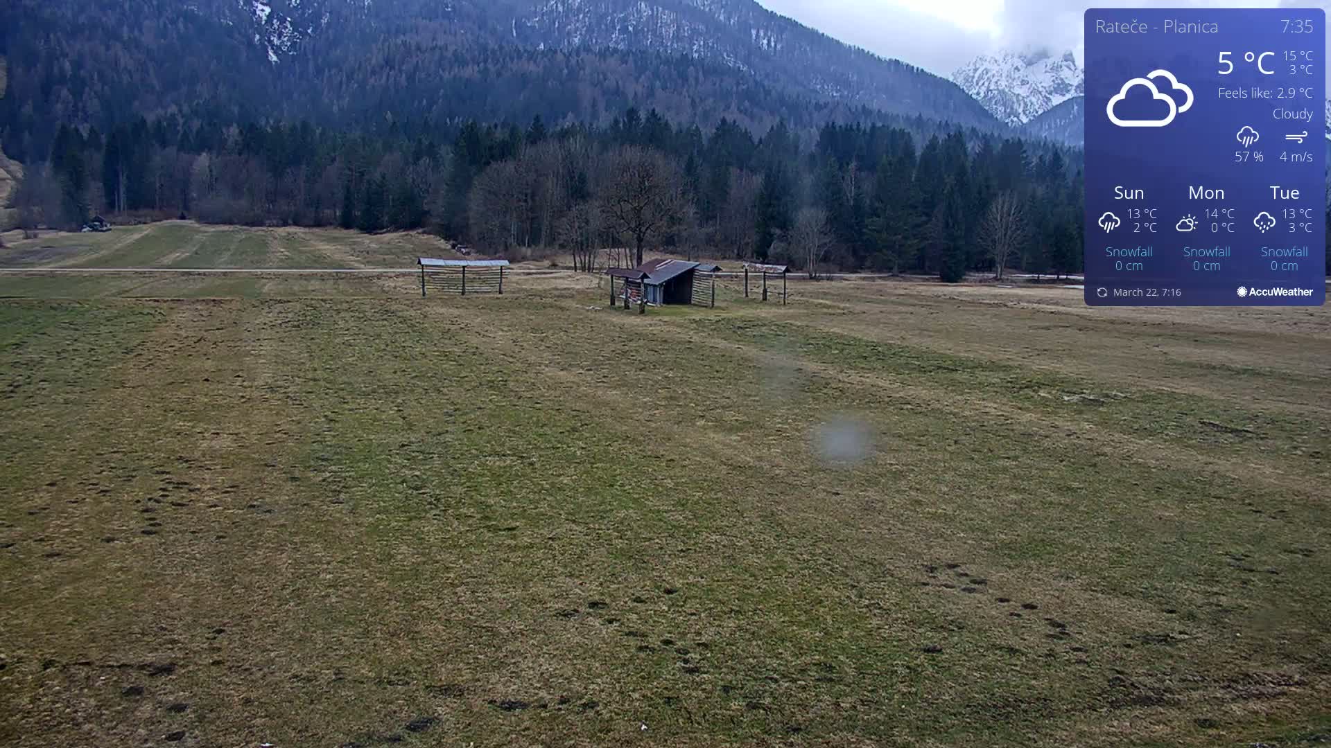 A cloudy, 5°C day reveals a flat, grassy field with a small shed and wooden structures, backed by a snow-capped mountain range and a dark evergreen forest.