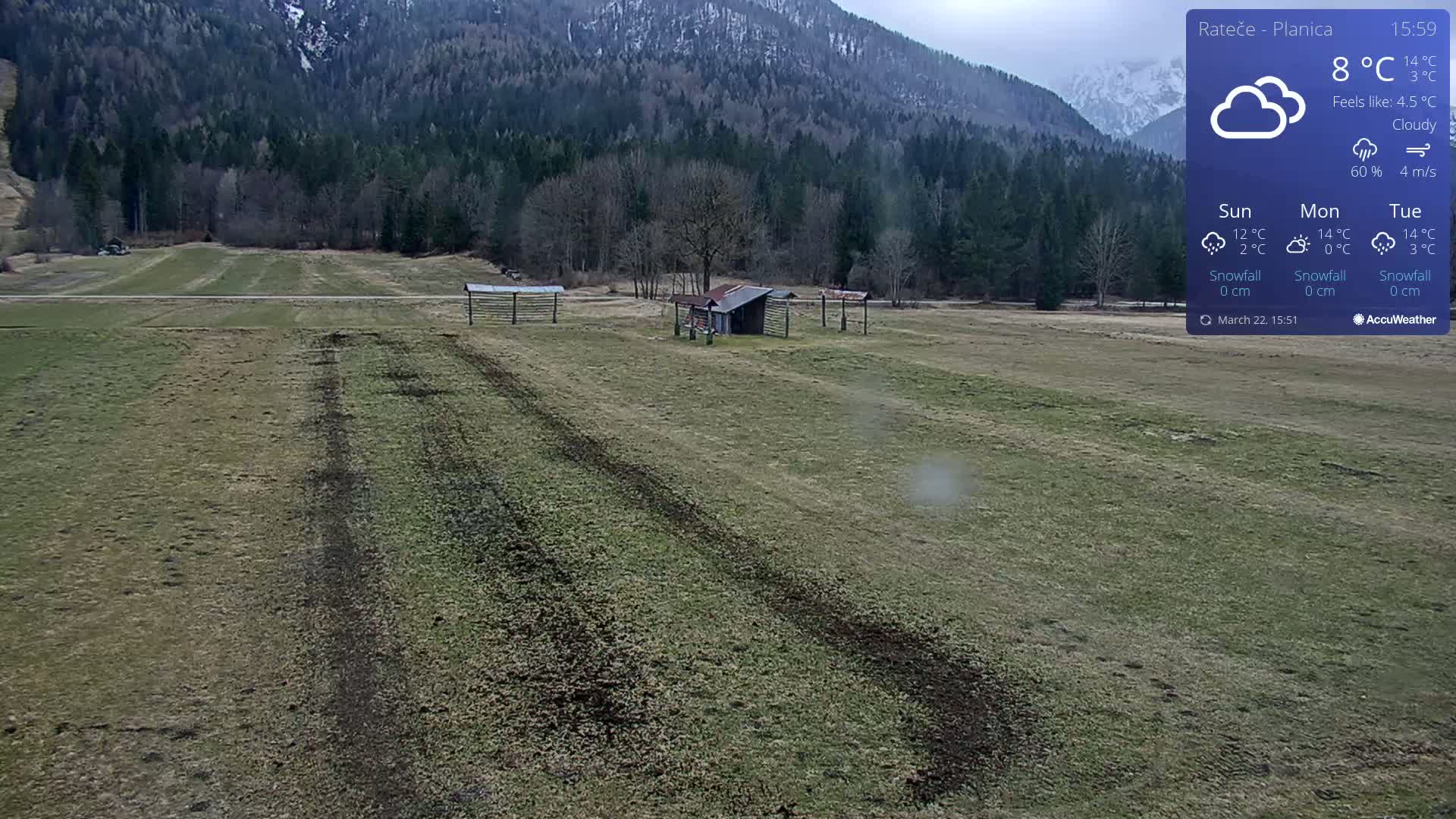 A cloudy, 8°C day shows a flat, grassy field with tire tracks leading to a small shed and wooden structures, all set against a backdrop of a mountain range.