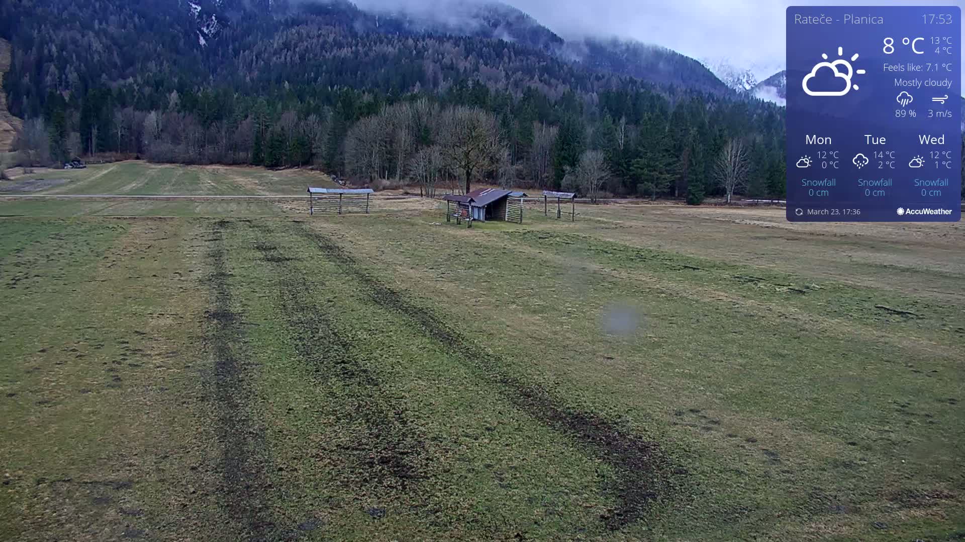 A mostly cloudy day shows a flat, grassy field with tire tracks, a small shed, and wooden structures, all set against a backdrop of a mountain range covered in a forest.