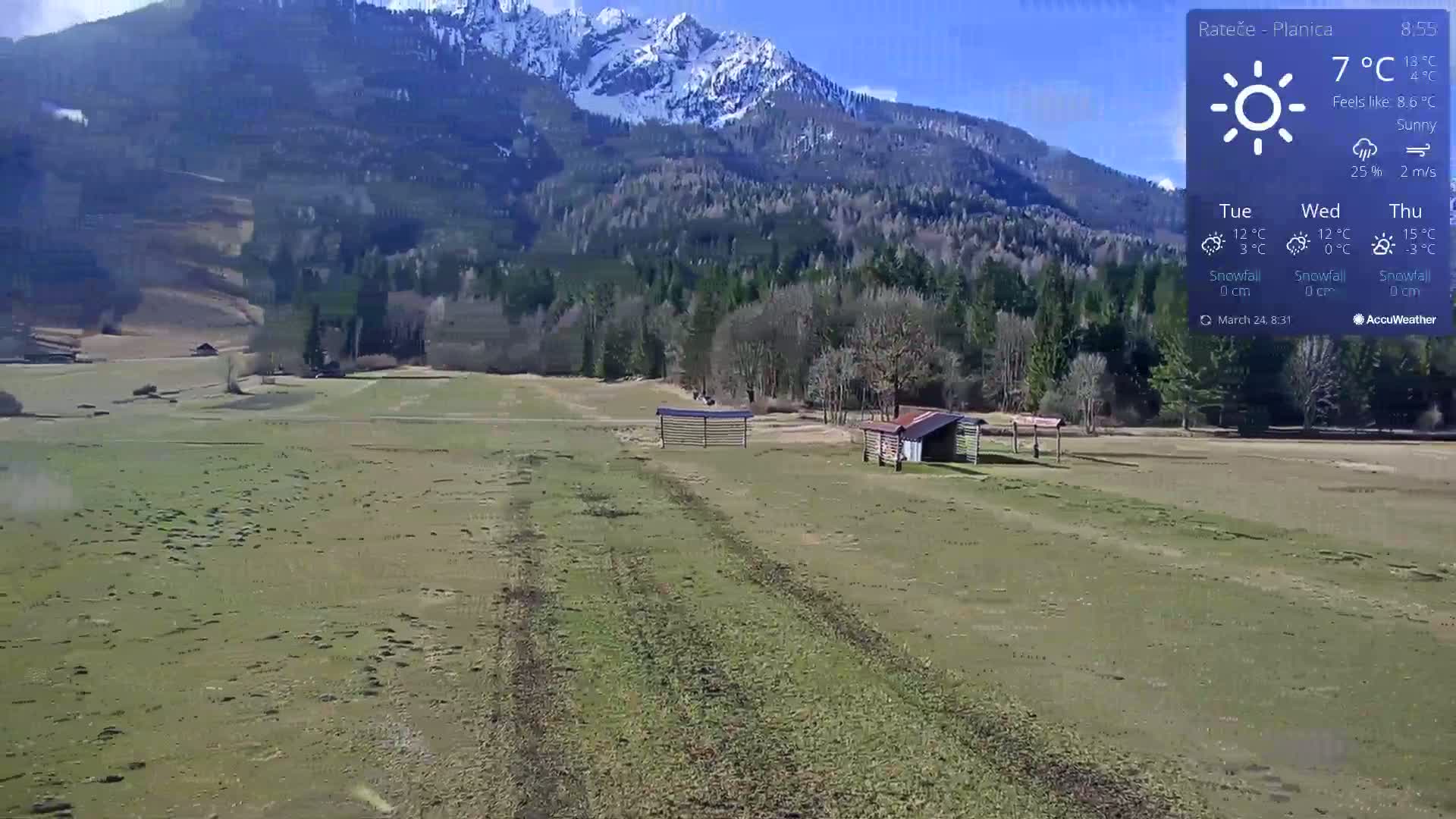 A sunny, mostly flat grassy field with tire tracks and two small structures is situated in front of a snow-capped mountain range and a forest.