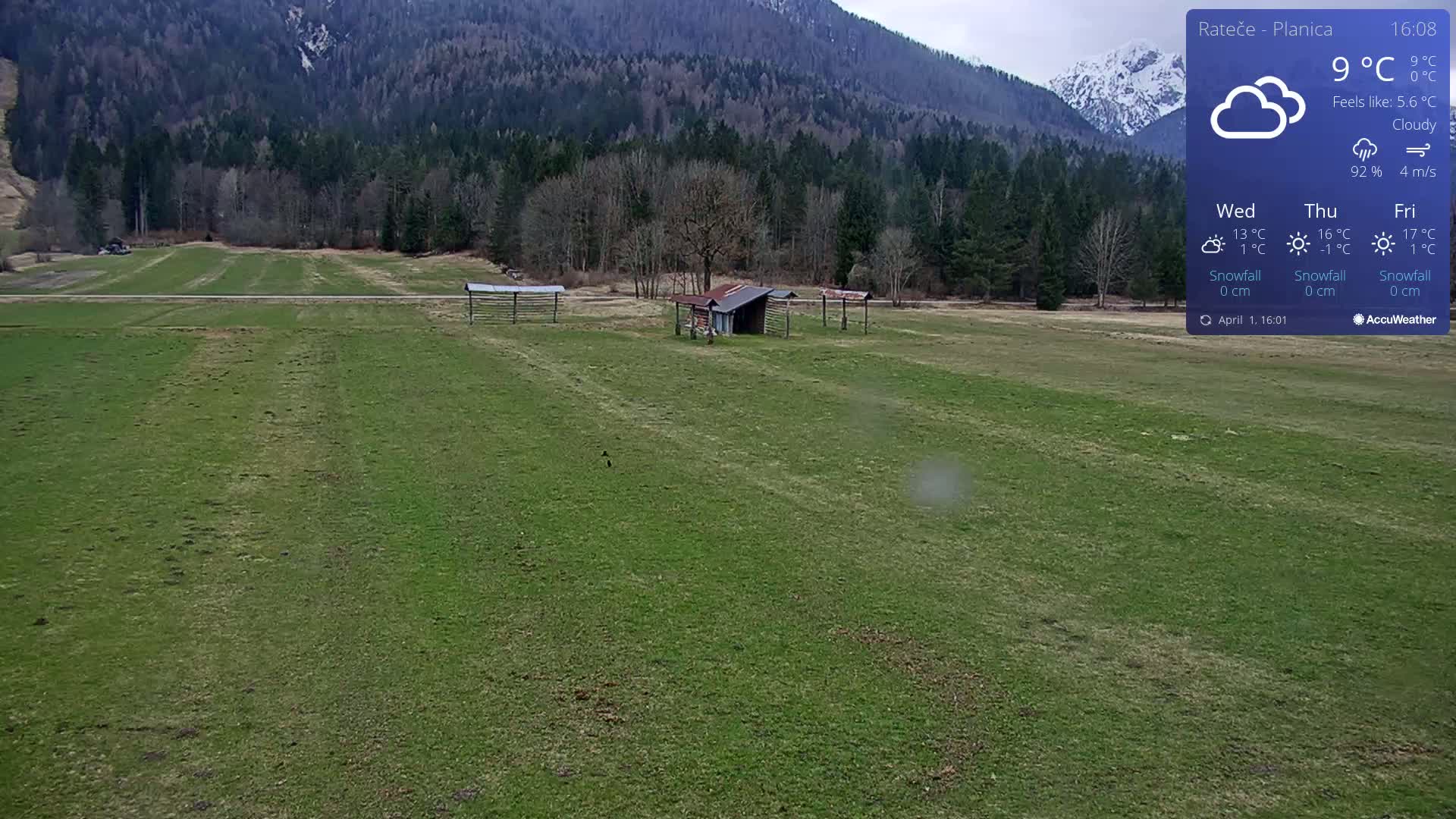 A green field with a small shed and three wooden structures stretches out beneath a cloudy sky in front of a mountain range.