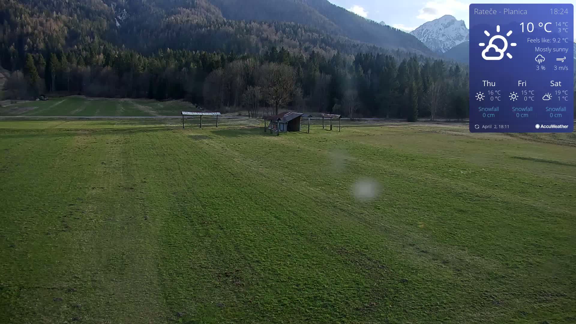 A mostly sunny, green field with a small shed and several open-air structures is nestled in a valley surrounded by a mountain range.