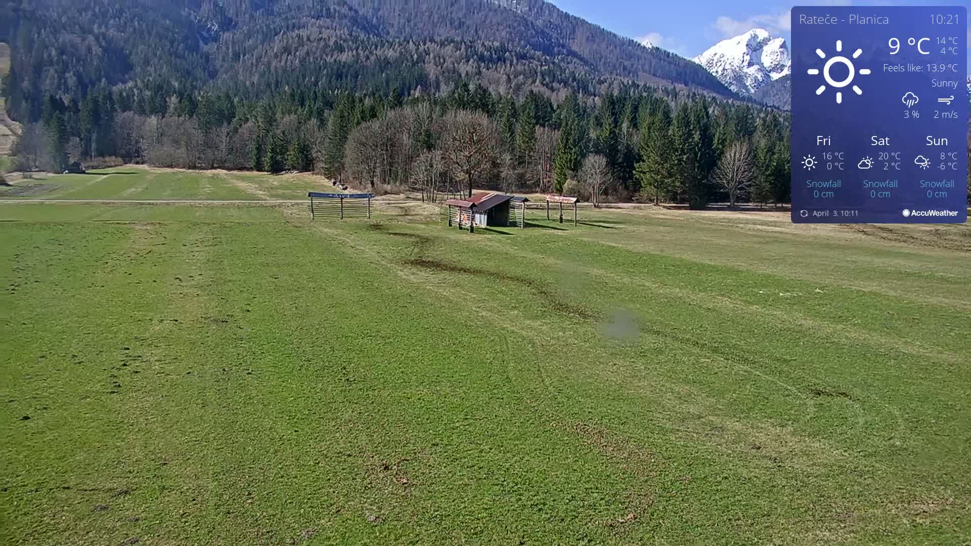 A sunny day reveals a grassy field with several small structures and a background of a forested mountain range under a clear sky.