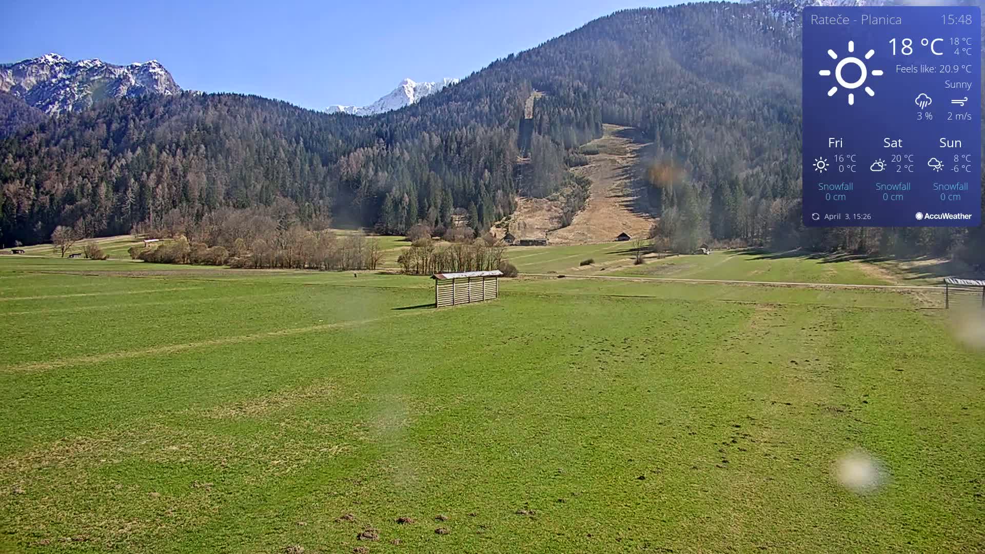 A sunny, expansive green field stretches out before snow-capped mountains under a clear blue sky.