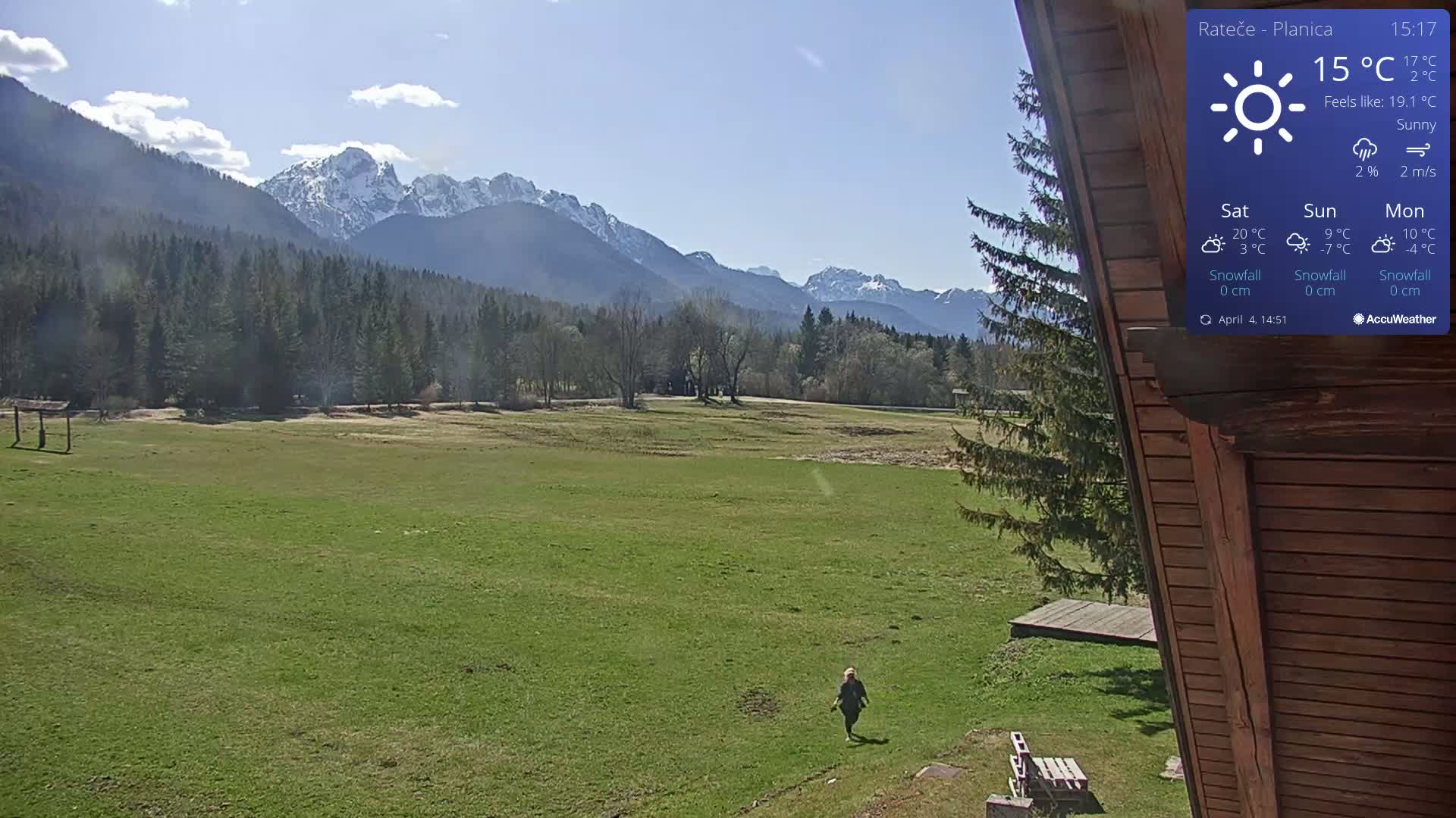 A person walks across a grassy field, with a mountain range in the background under sunny conditions.