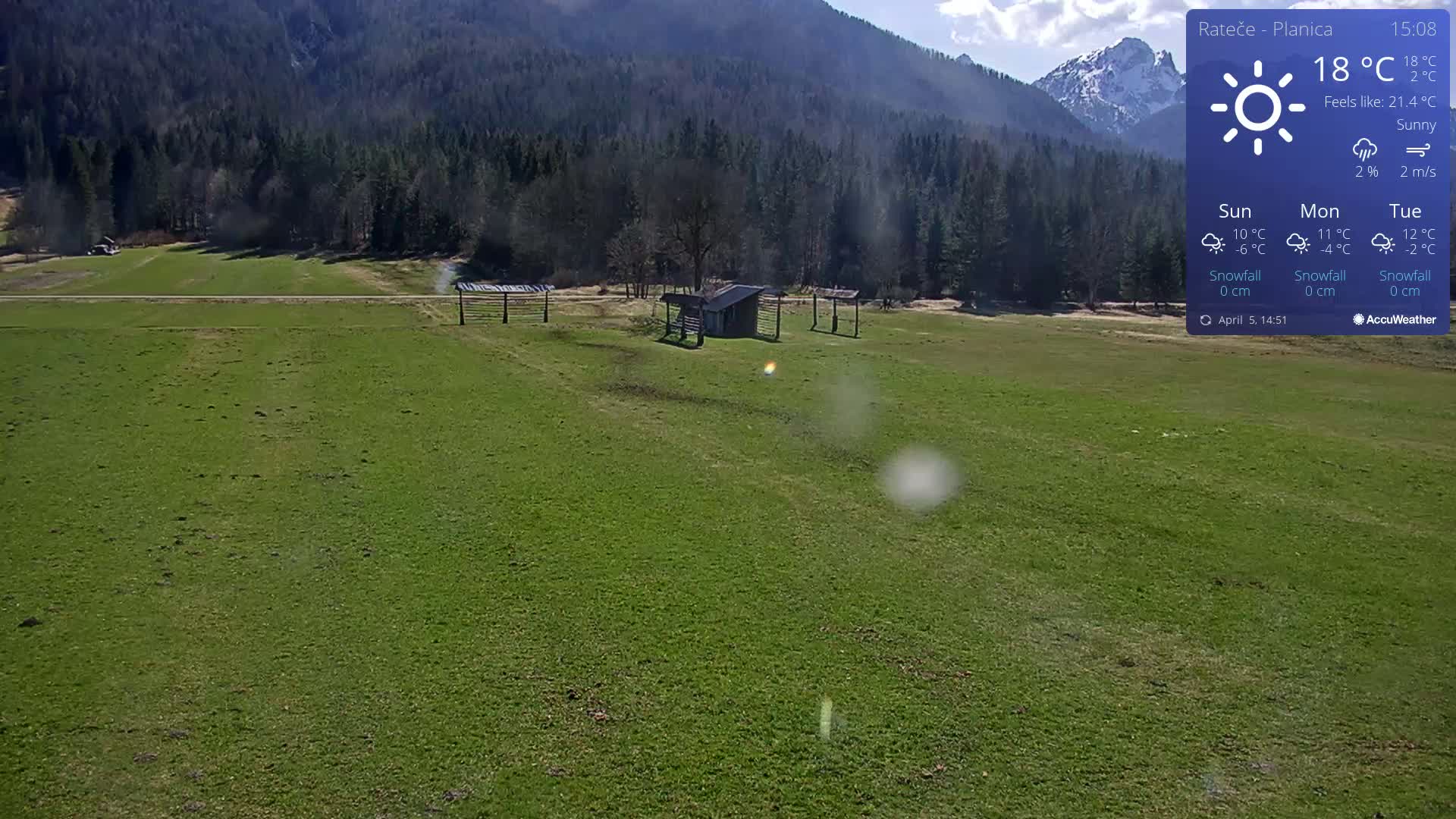 A sunny day in a green field with a small shed and wooden structures, backed by a forest and mountains.