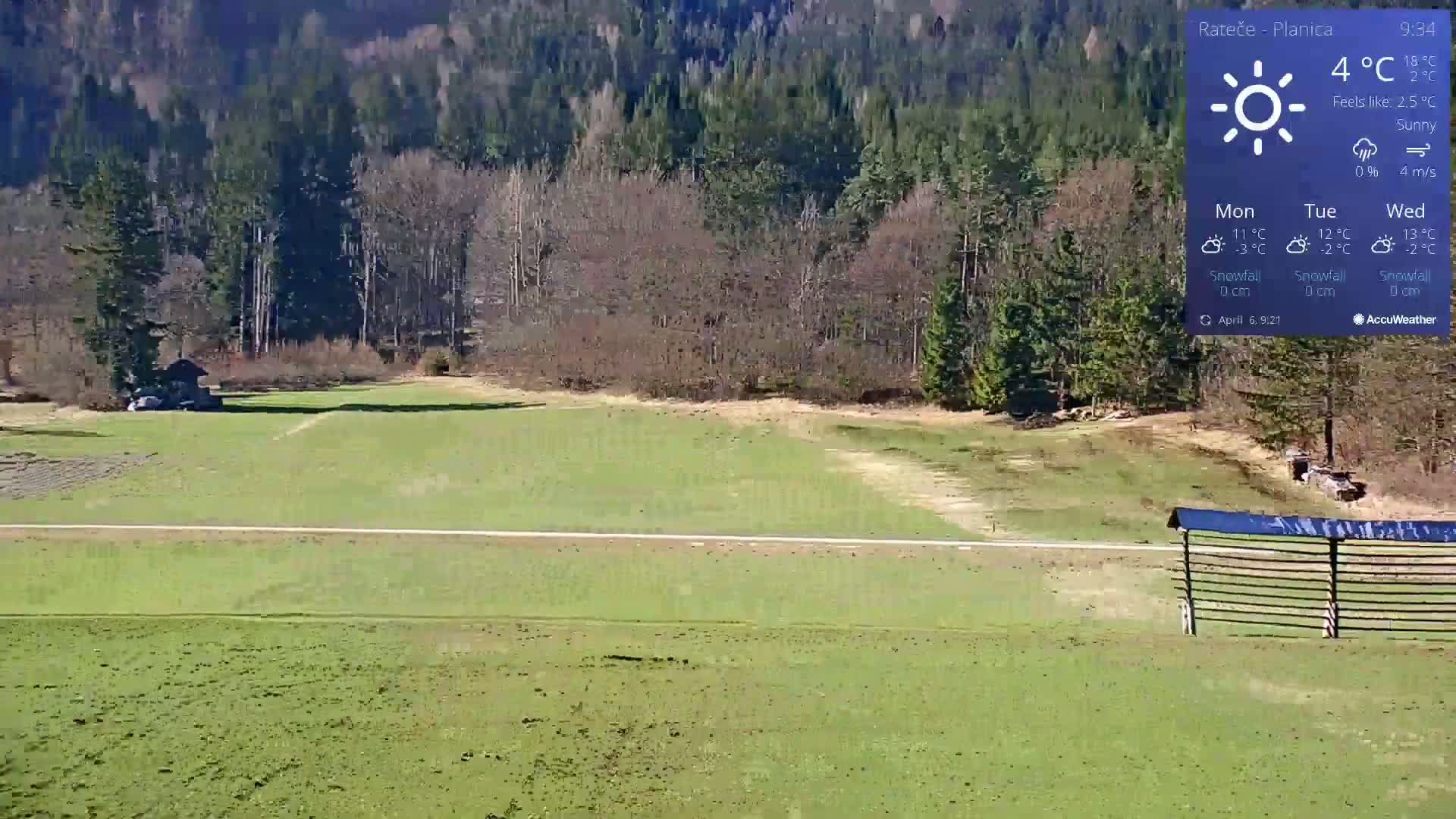 A sunny, grassy field with a small building in the distance, bordered by a forest, and a dark-colored barrier in the foreground.