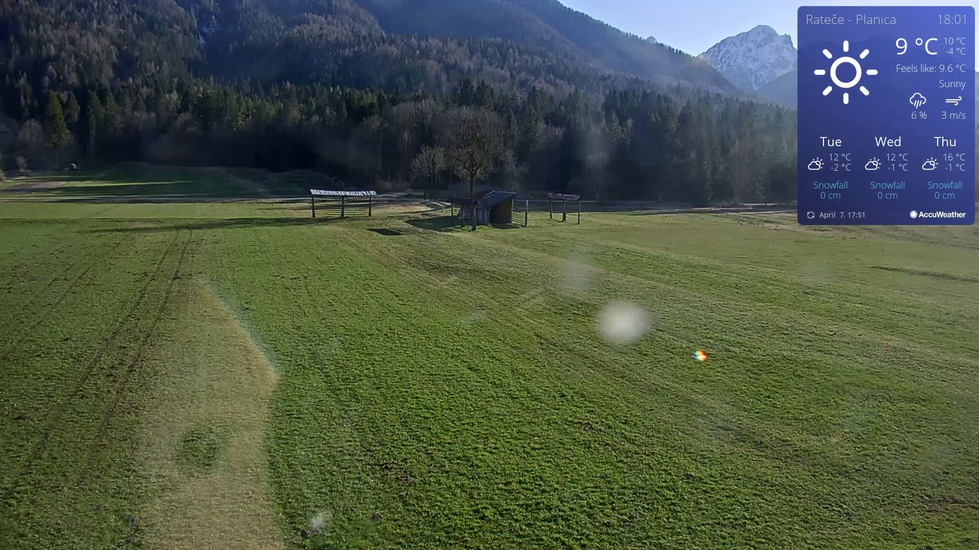A sunny, expansive green field with a small wooden structure and simple shelters sits below a background of dark green forested hills and snow-capped mountains.