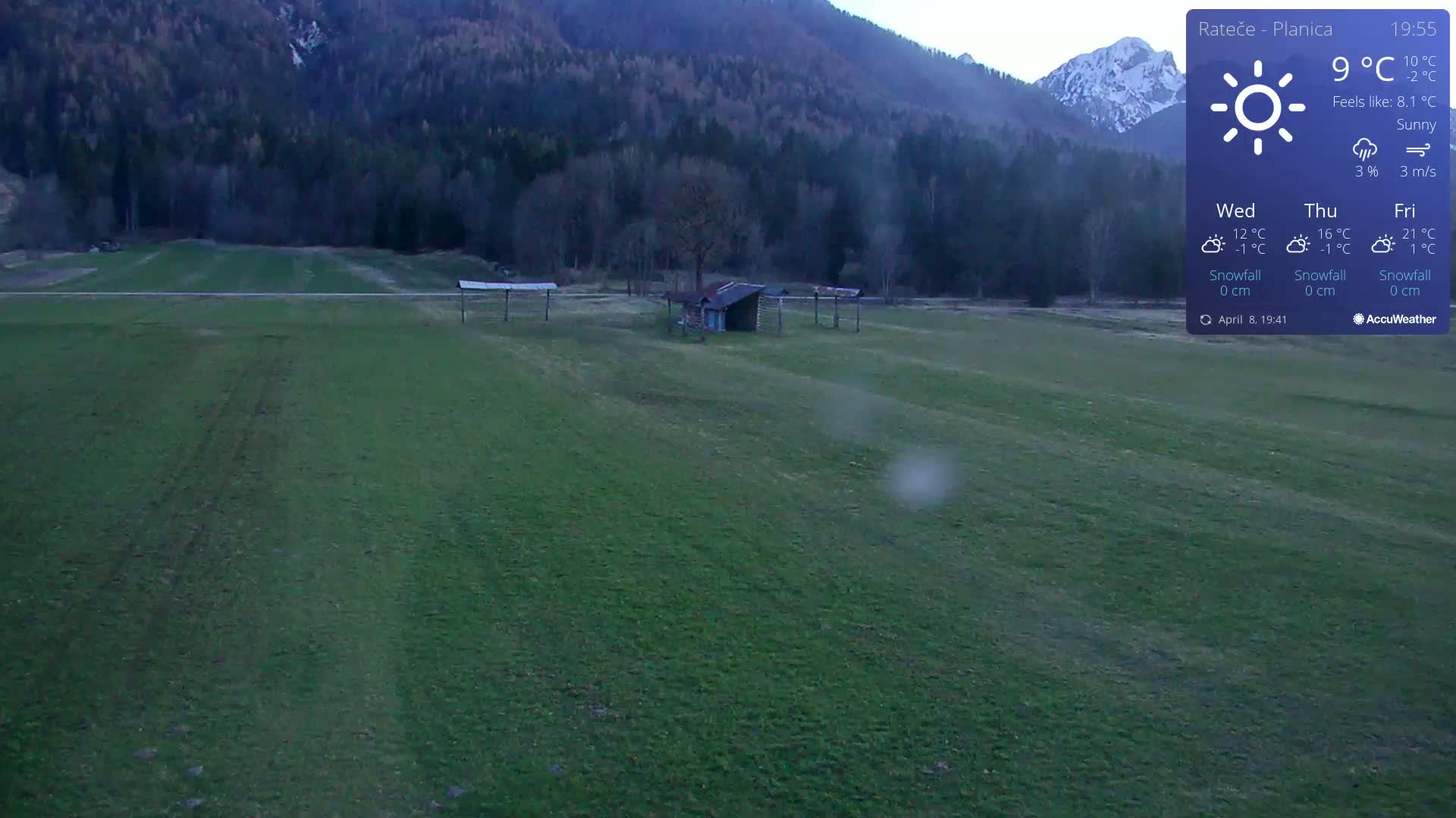 A sunny day reveals a grassy field with a small shed and several open-air structures, backed by a mountain range under a mostly clear sky.