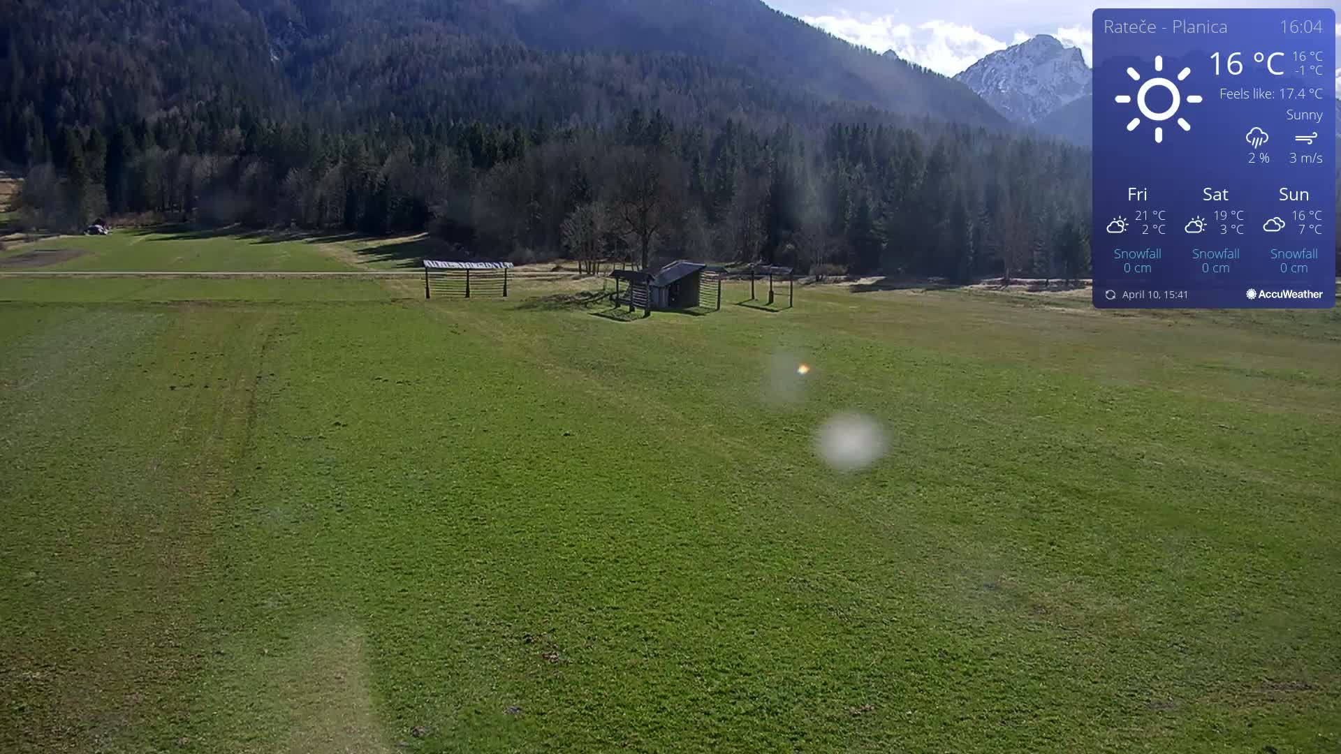 A sunny day reveals a verdant field, a small shed, and some wooden structures, all set against a backdrop of forested mountains.