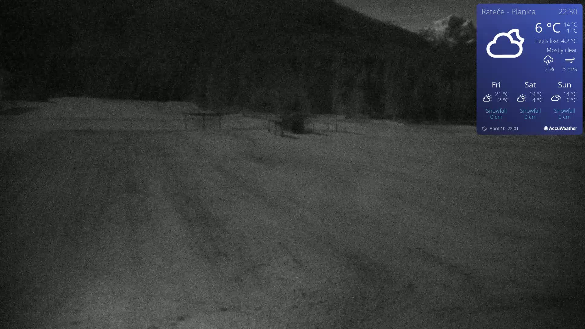 A mostly clear night scene shows a snow-covered, flat area with a dark background of trees and a mountain under a mostly clear sky.
